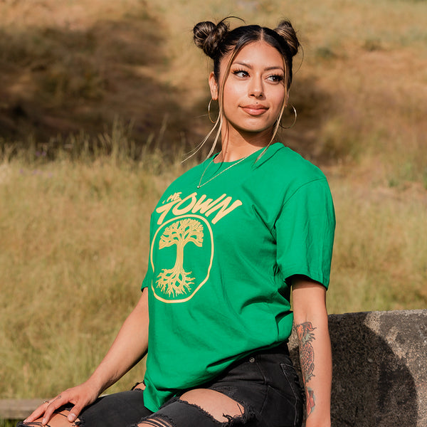 Woman smiling wearing kelly tee with golden yellw print with words The Town and underneath that a circle with the Oaklandish tree inside.