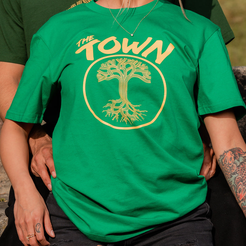 Close up shot of person wearing a green tee with The Town and underneath that a circle with the Oaklandish tree inside, in golden yellow ink.