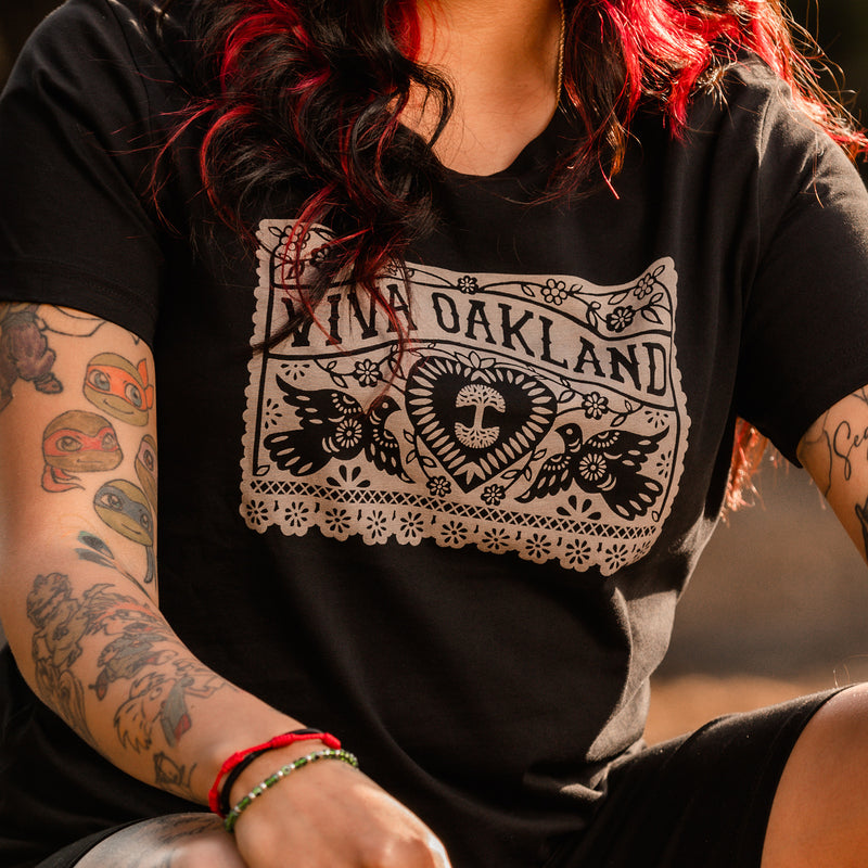 Closeup image of chest area of woman wearing Viva Oakland design featuring paper cut out style Mexican-inspired design with heart and Oaklandish tree and birds and the words Viva Oakland.