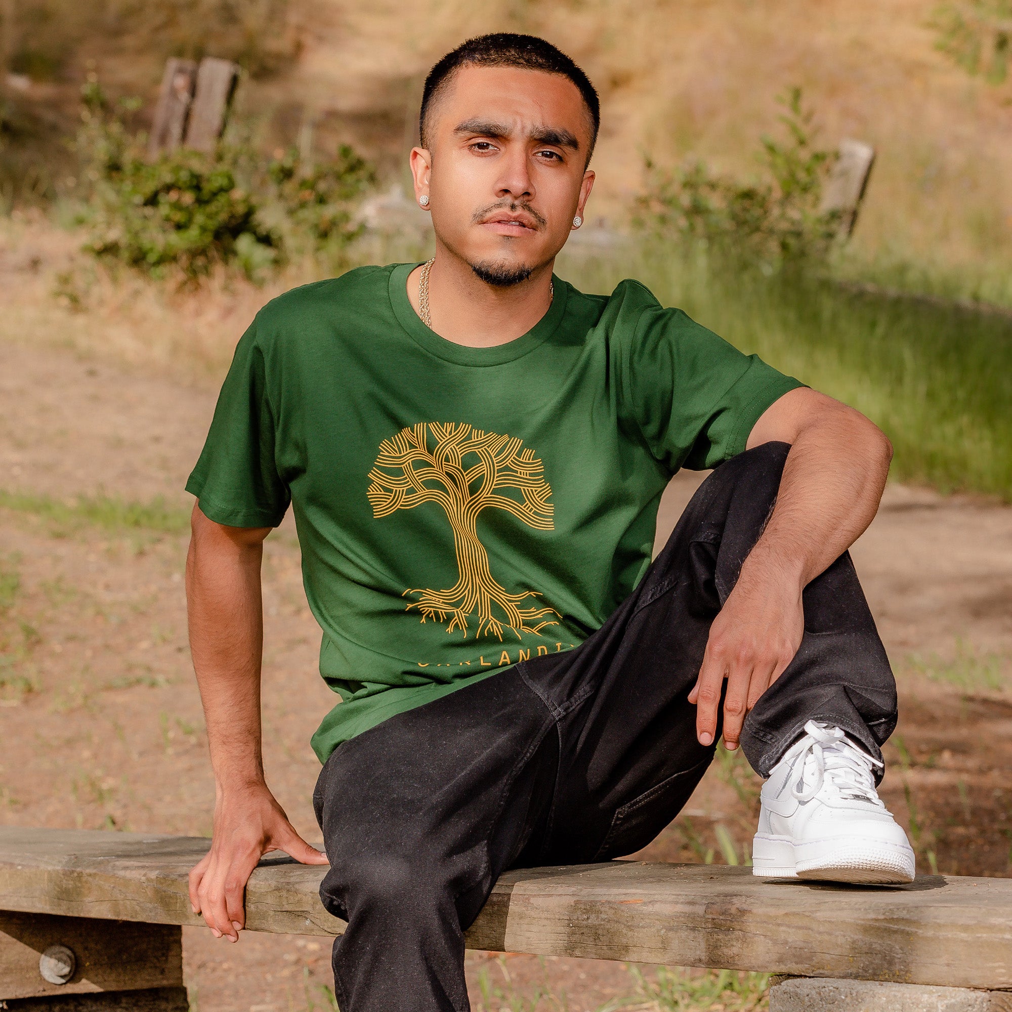 Male model seated on bench outdoors with one foot up wearing forest green tee with Oaklandish tree logo in golden yellow on front of tee.