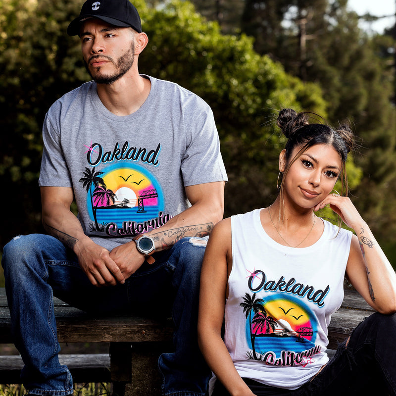 Male and female model seated outdoors wearing grey tee and white tank top with multicolor bright ink colors coastal bay scene with palm trees and bridges and hearts, text spelling Oakland, California.