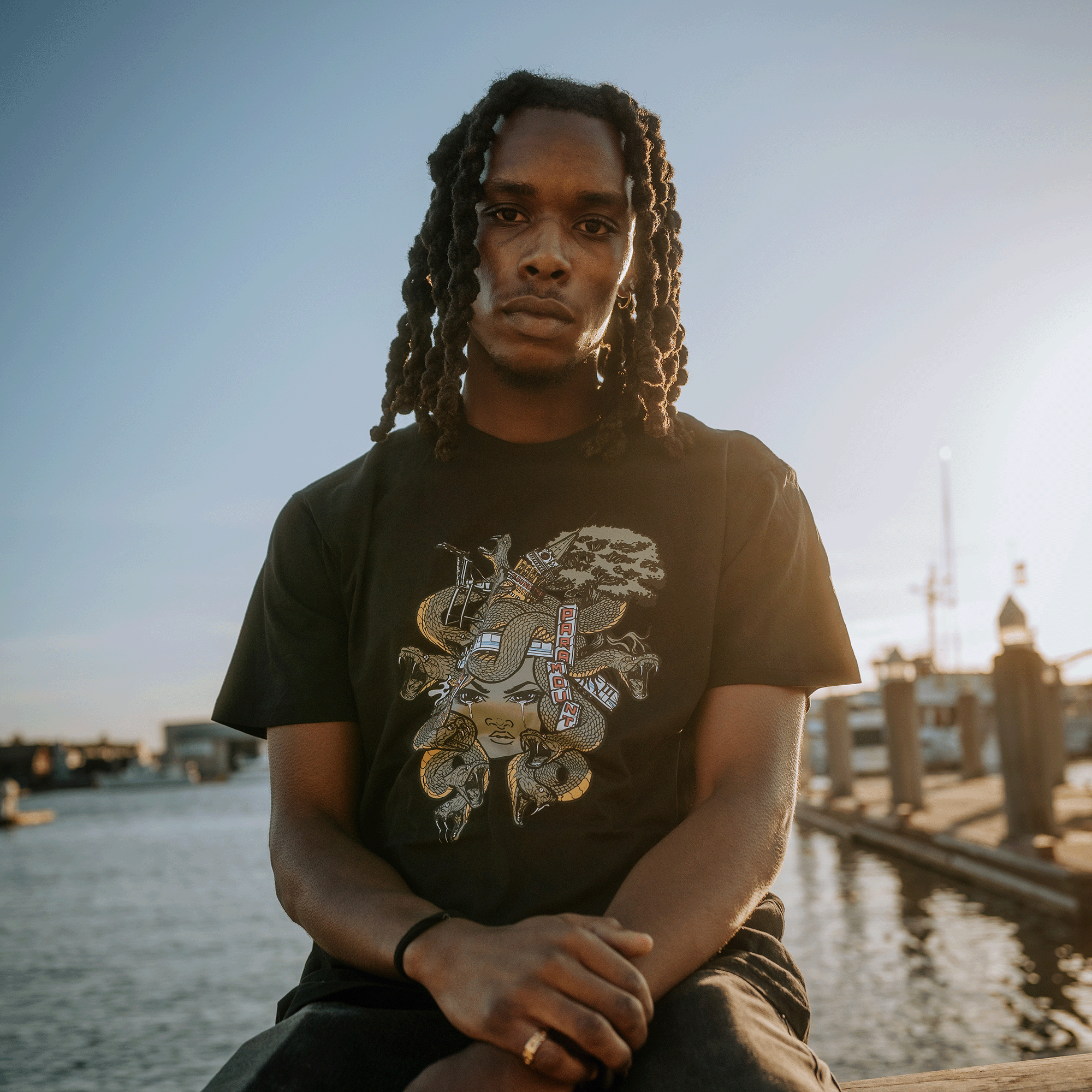 Model wearing a black t-shirt with elaborate Medusa monster of the mind graphic while sitting on dock.