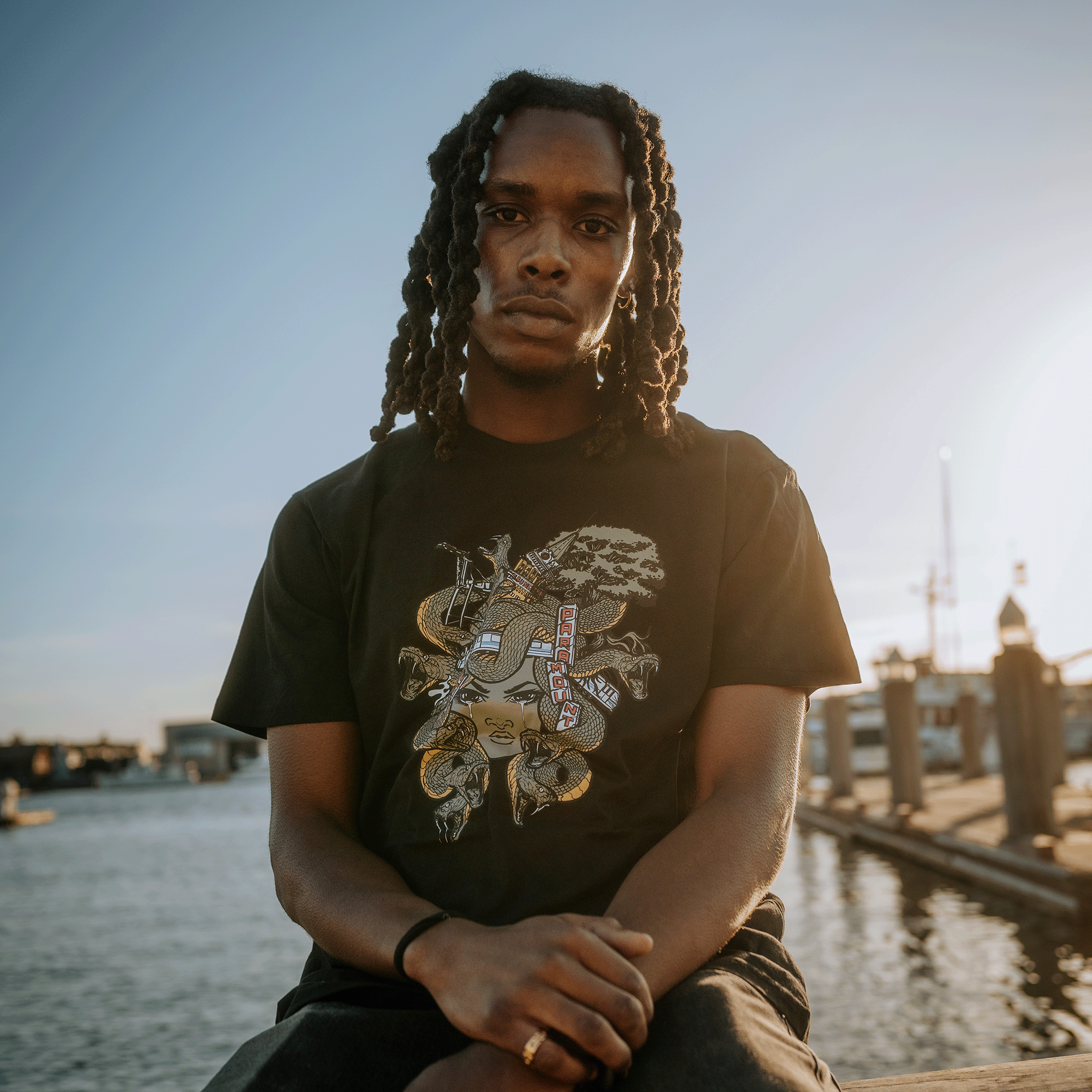 A person with long braided hair sits on a ledge near a waterfront, wearing the Medusa Tee from Oaklandish's men's collection. The black t-shirt, made of 100% cotton and featuring intricate artistic designs, glows warmly in the setting sun, illuminating the calm water and docked boats as the person looks contemplative.