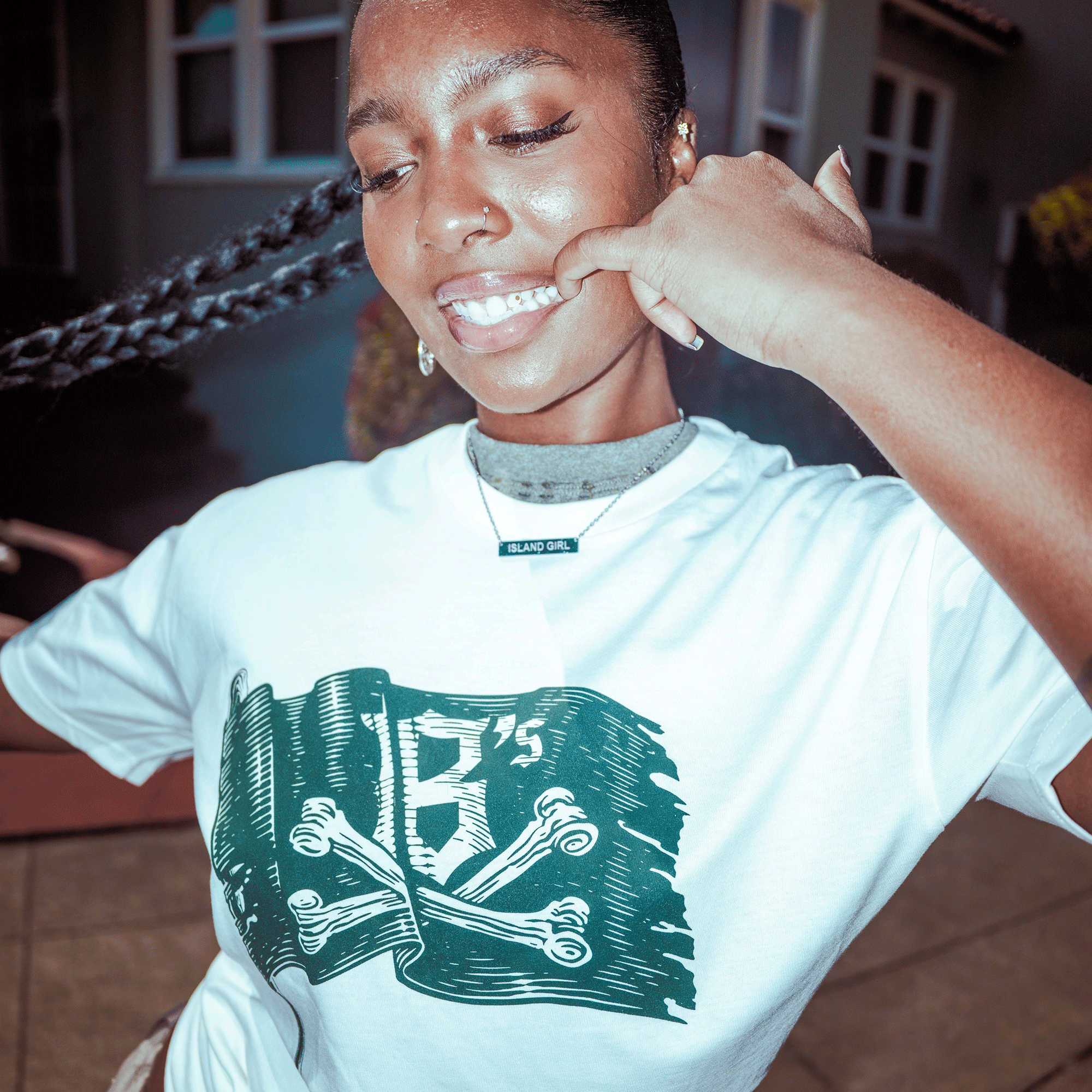 A young woman smiles outdoors, tugging her cheek while wearing the Oakland Ballers Jolly Roger Tee—a white shirt with a dark green crossed bones graphic and "18’s" text, showing love for Oakland's Pioneer League pro baseball team. Her necklace reads "ISLAND GIRL.