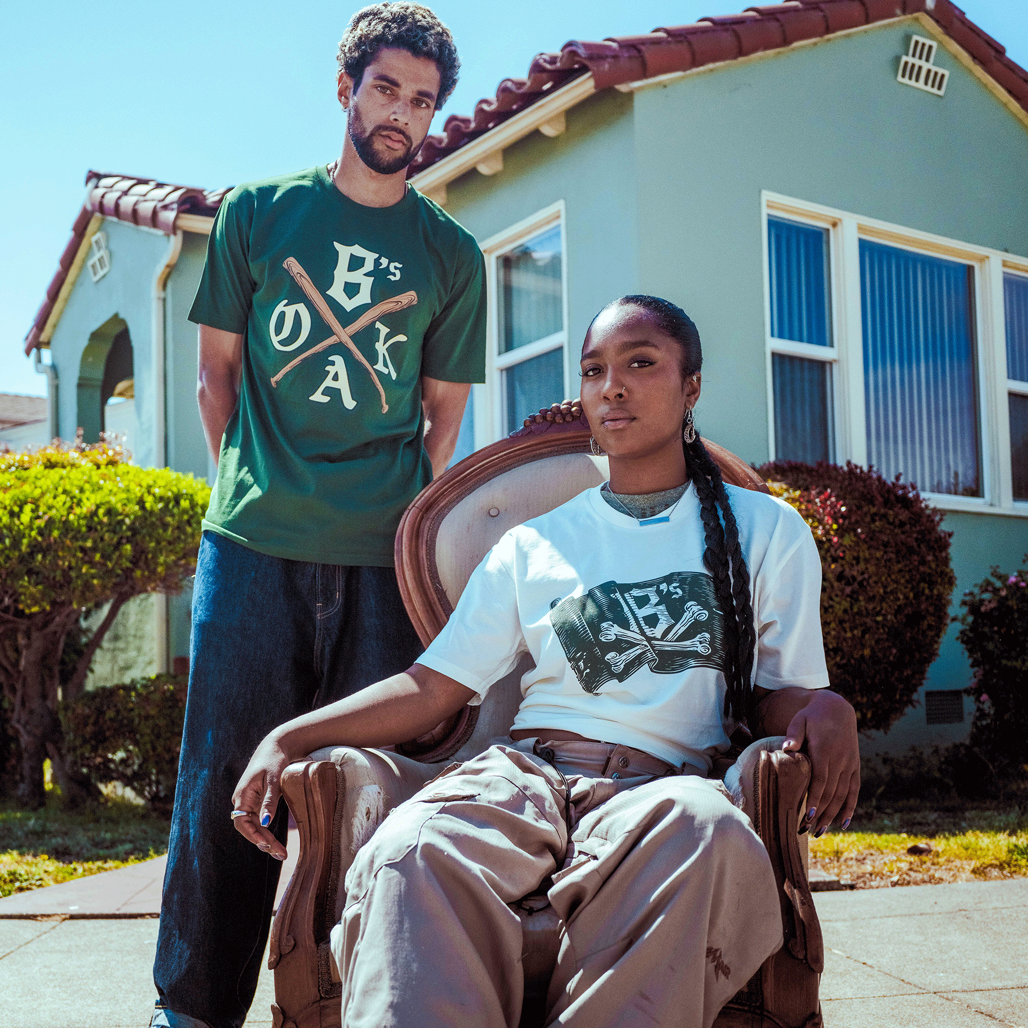 A man stands behind a seated woman outdoors before a light-green house. He wears the Oakland Ballers OAK Bat Tee; she sits confidently, both facing the camera under bright sunlight.