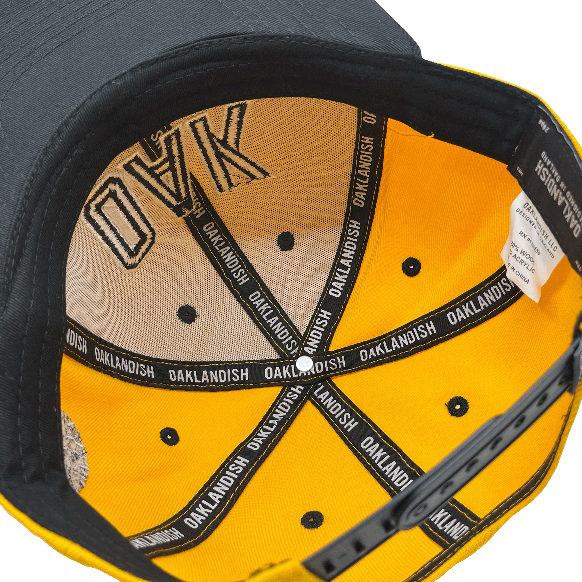 The image shows the inside of an OAK Block Snapback by Oaklandish, which has a black exterior and yellow lining. The headwear features "OAK" written on the inner part of the brown headband, and its seams repeatedly display "OAKLANDISH" in white text on black fabric strips. Eyelets are visible around the inner crown.