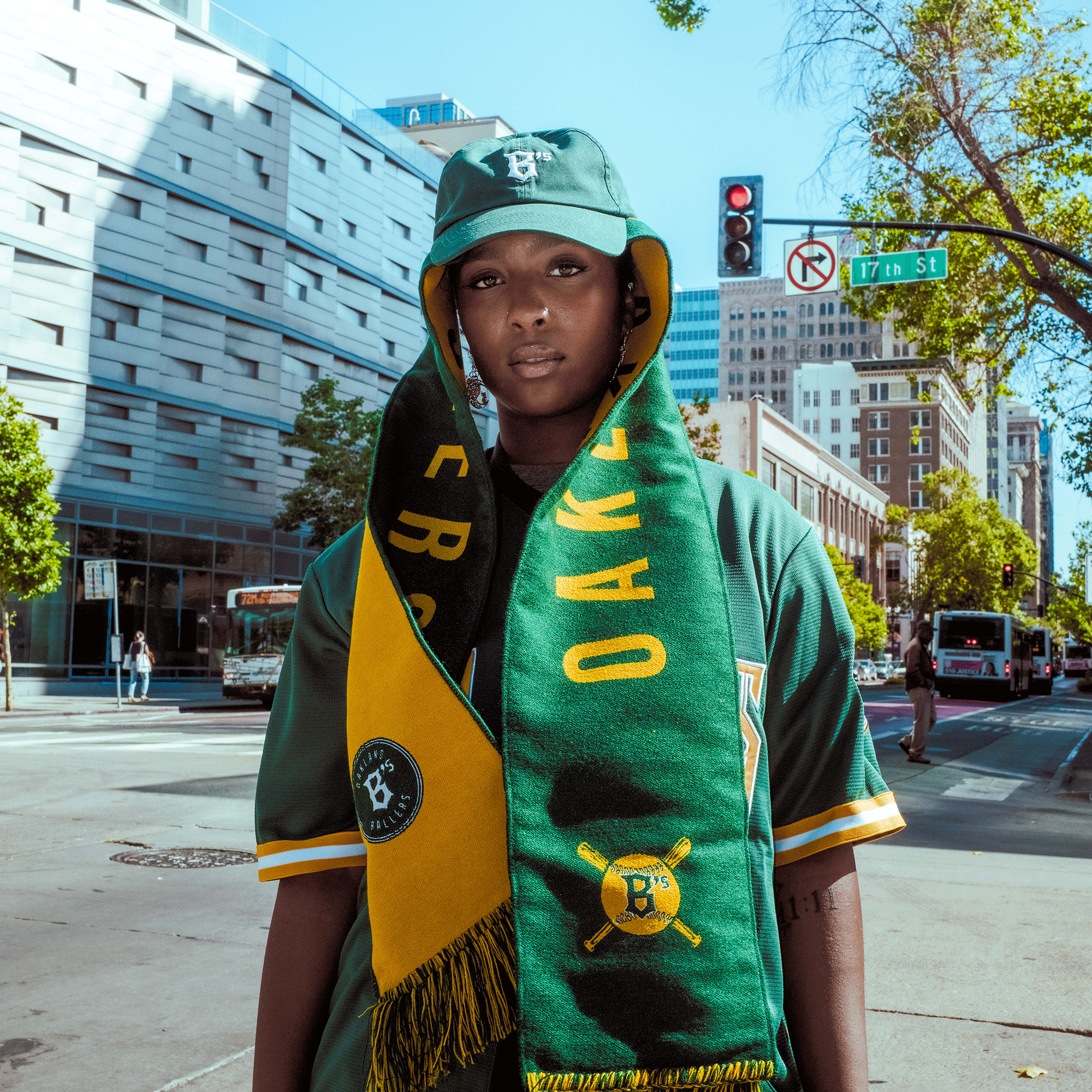 A person stands on a city street in Oakland Ballers gear, including a green jersey, bucket hat, and an Oakland Ballers Scarf. A “17th St” sign highlights the home of Oakland’s pro teams, including the new Pioneer League club.