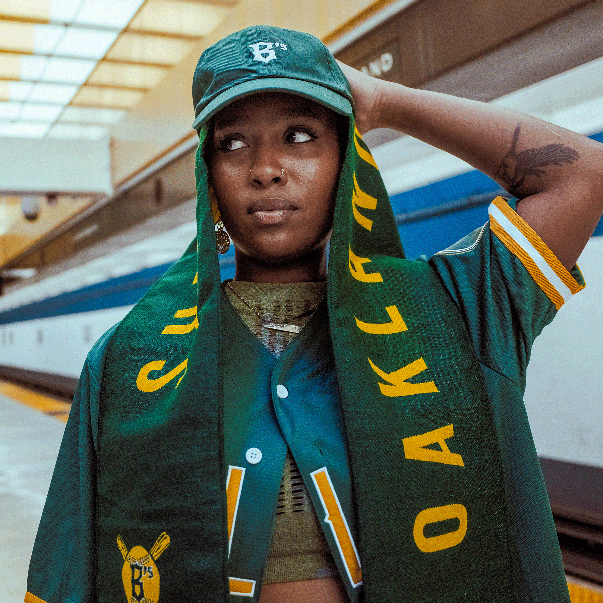 A woman at a train platform, proudly wearing an Oakland Ballers Scarf by Oakland Ballers, a green cap, and jersey, shows her city pride—celebrating the new Pioneer League pro baseball team: the Oakland Ballers.