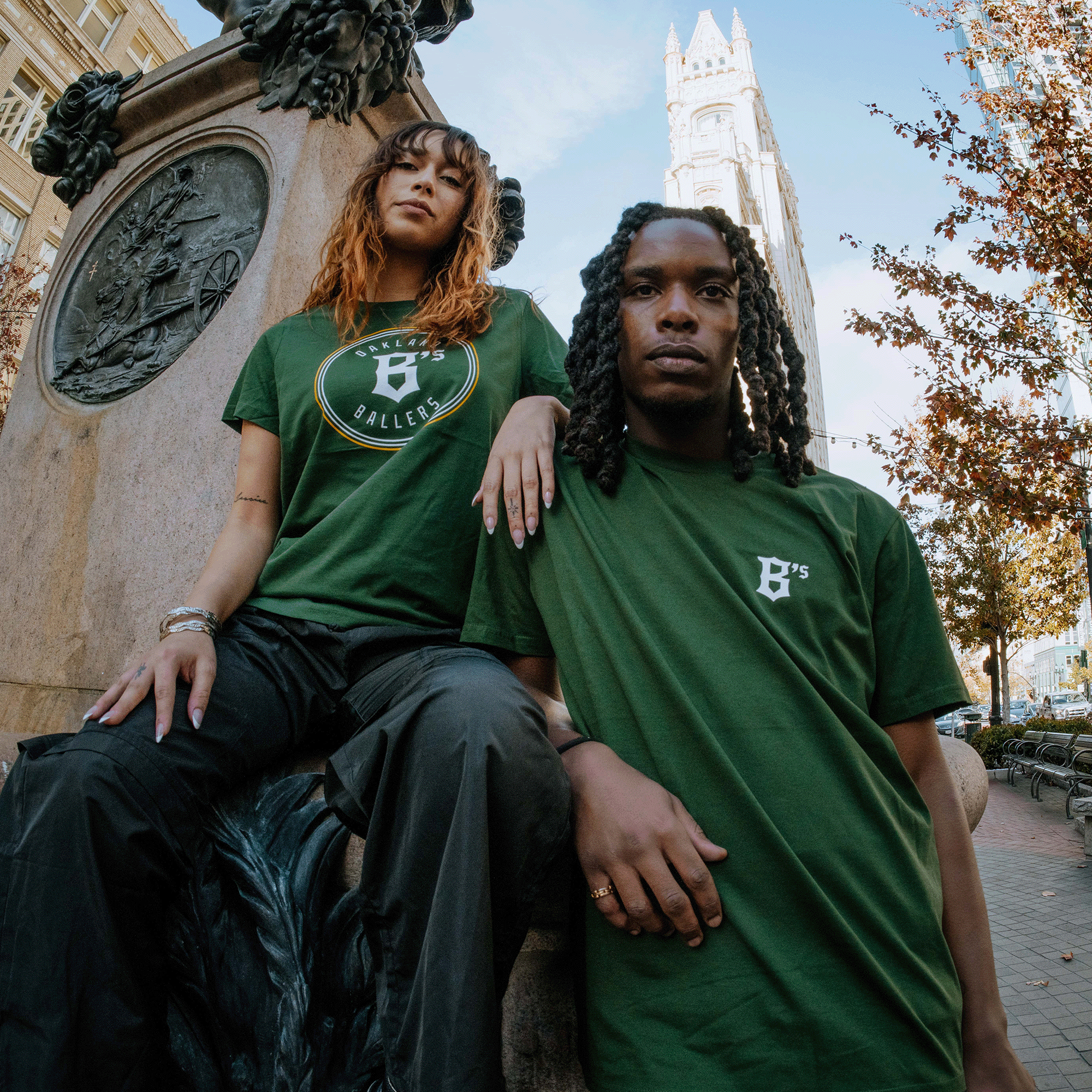 Two individuals pose in front of a city monument. The person on the left is seated with a hand on the monument, wearing an Oakland Ballers Logo Tee and black pants, and has curly hair. The person on the right stands, also in a green T-shirt with "B's" logo and long braided hair. A tall building and autumn trees create a picturesque backdrop evocative.