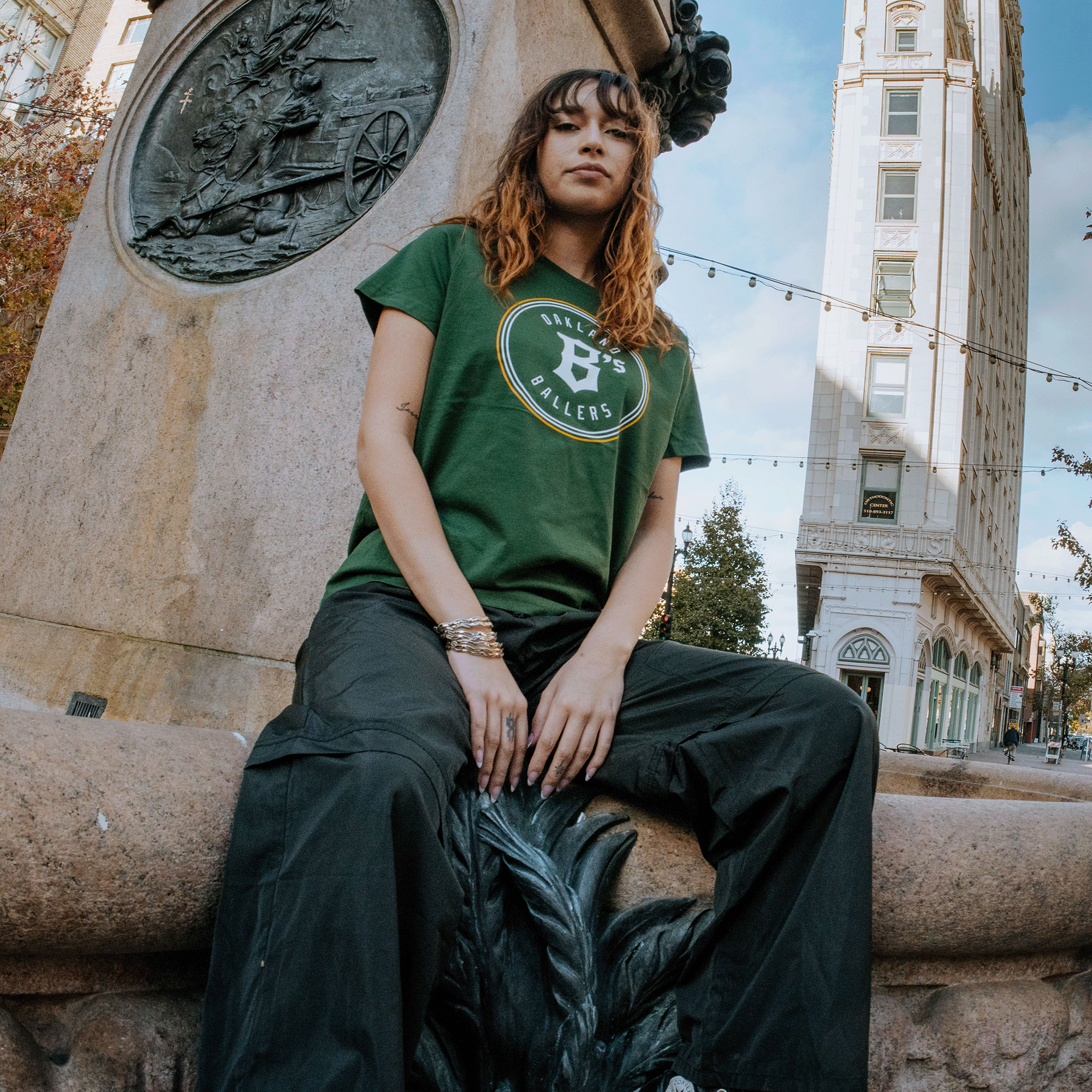 A person with wavy hair sits on a stone structure in an urban setting, dressed in a Women's Oakland Ballers Logo Tee and black pants. The green t-shirt, branded by Oakland Ballers, showcases a circular design with text on the front. Behind them is a tall building, trees, and string lights hanging across the street, creating a lively city atmosphere.