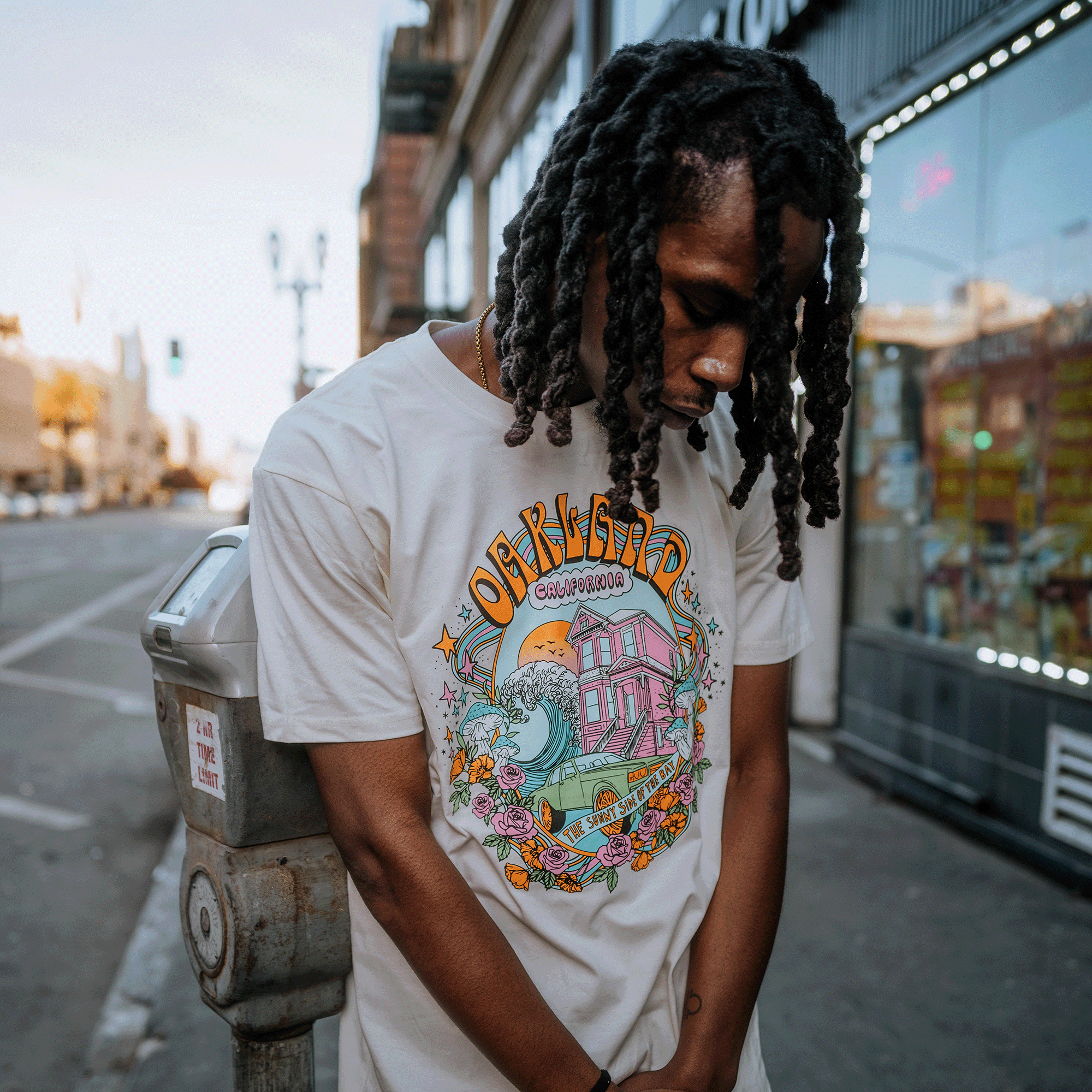 A person with long, dark dreadlocks stands leaning against a parking meter on a city street. They are wearing a vibrant Oakland Dream Tee by Oaklandish, made from 100% cotton in a classic fit and adorned with whimsical graphics depicting a house with fireworks and flowers. The urban background features buildings and a clear sky.
