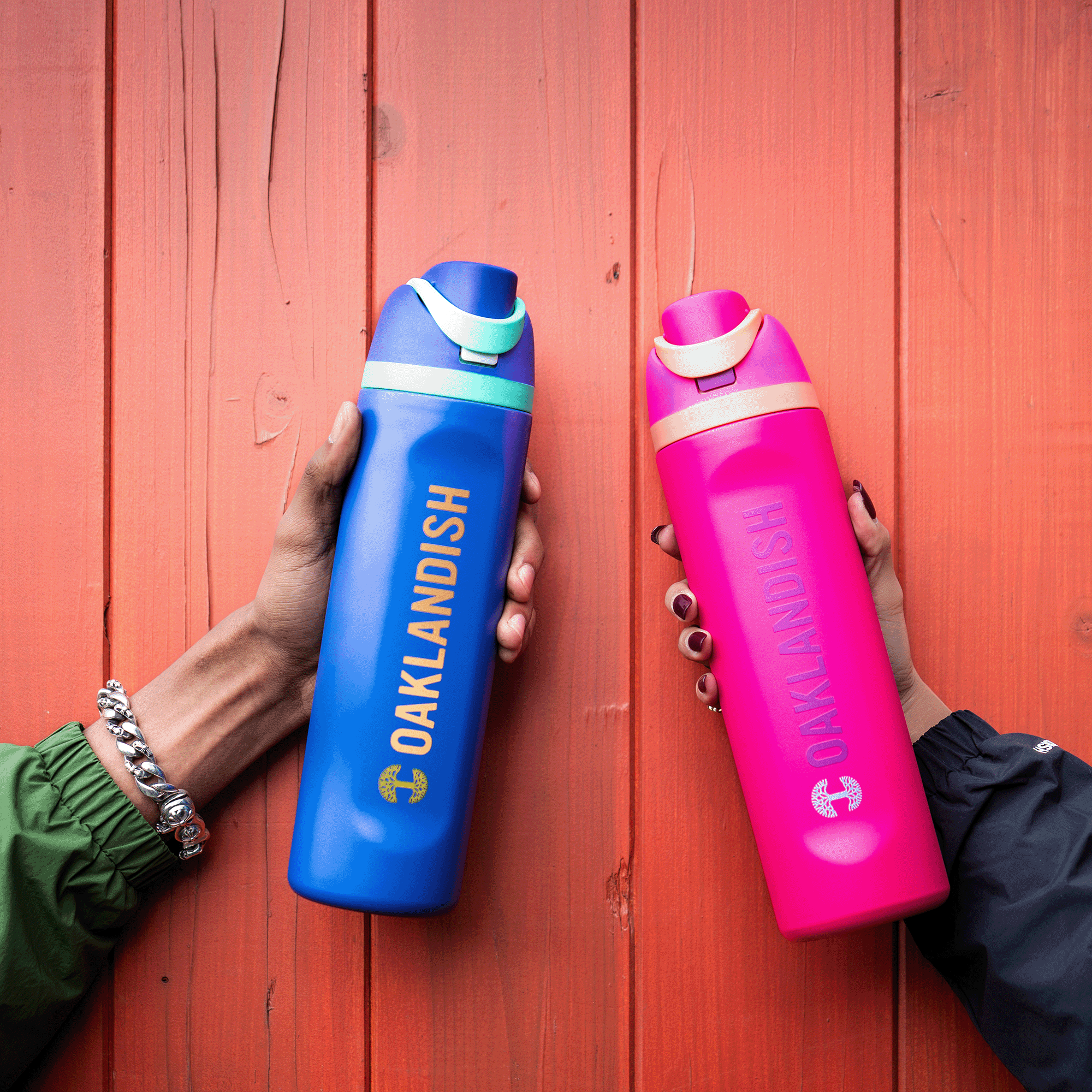 Two hands hold Oaklandish Colorful Bottles against an orange wooden background—one blue with "OAKLANDISH" in yellow, the other pink with "OAKLANDISH" in lighter pink. Both feature the Oaklandish tree logo and flip-top lids.
