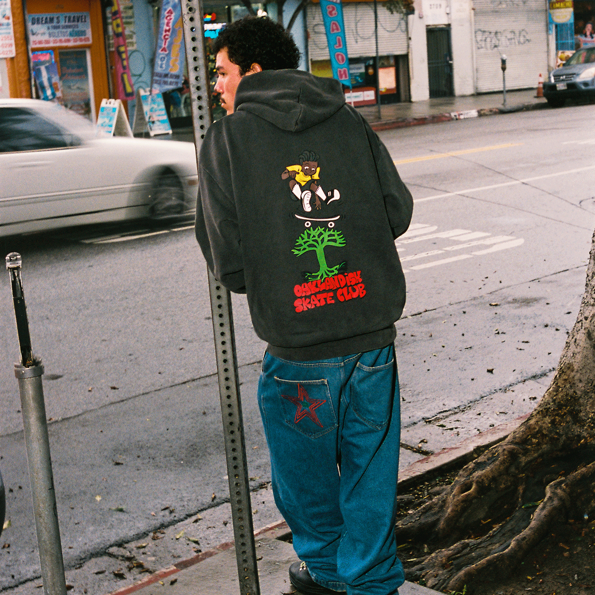 Wearing the Oaklandish Skate Club Hoodie, a person in blue jeans stands on a wet sidewalk by a tree. City shops, a street sign, and skateboard culture vibes appear on the overcast street. Brand: Oaklandish.