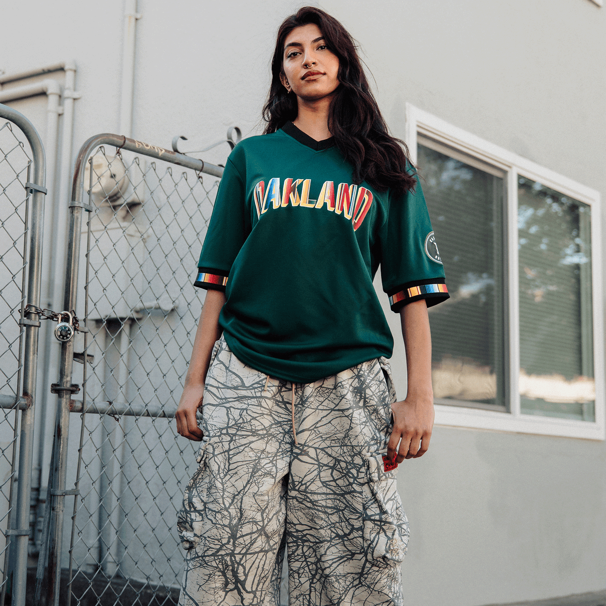 A person wears a dark green Peloteros Jersey with “OAKLAND” in bold, tri-colored letters and rainbow-striped cuffs, paired with baggy, light-colored pants featuring a dark, tree-branch-like pattern, posing outdoors in front of a chain-link fence.