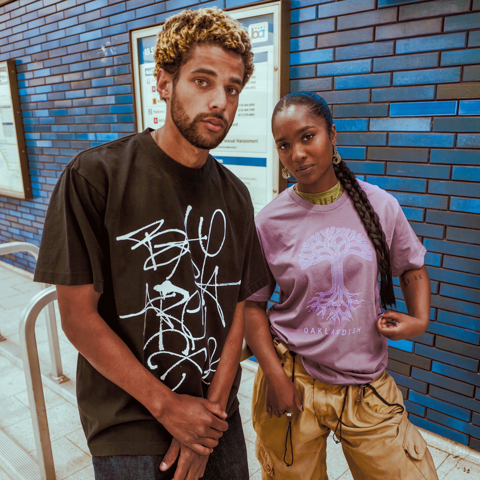 A man in a men's black heavy-weight cotton tee and a woman in a lavender Oaklandish Presto Tee with braided hair and tan cargo pants stand together by blue-tiled walls and transit maps.