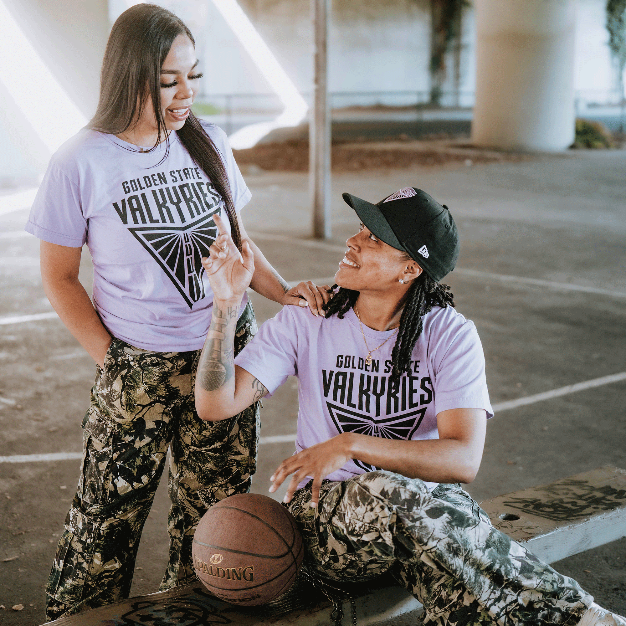 Two people stand on a basketball court under a bridge, both in matching GSV Golden State Valkyries Primary Premium Tees and camouflage pants. One sits with a basketball, smiling at the other, who stands nearby and smiles back.