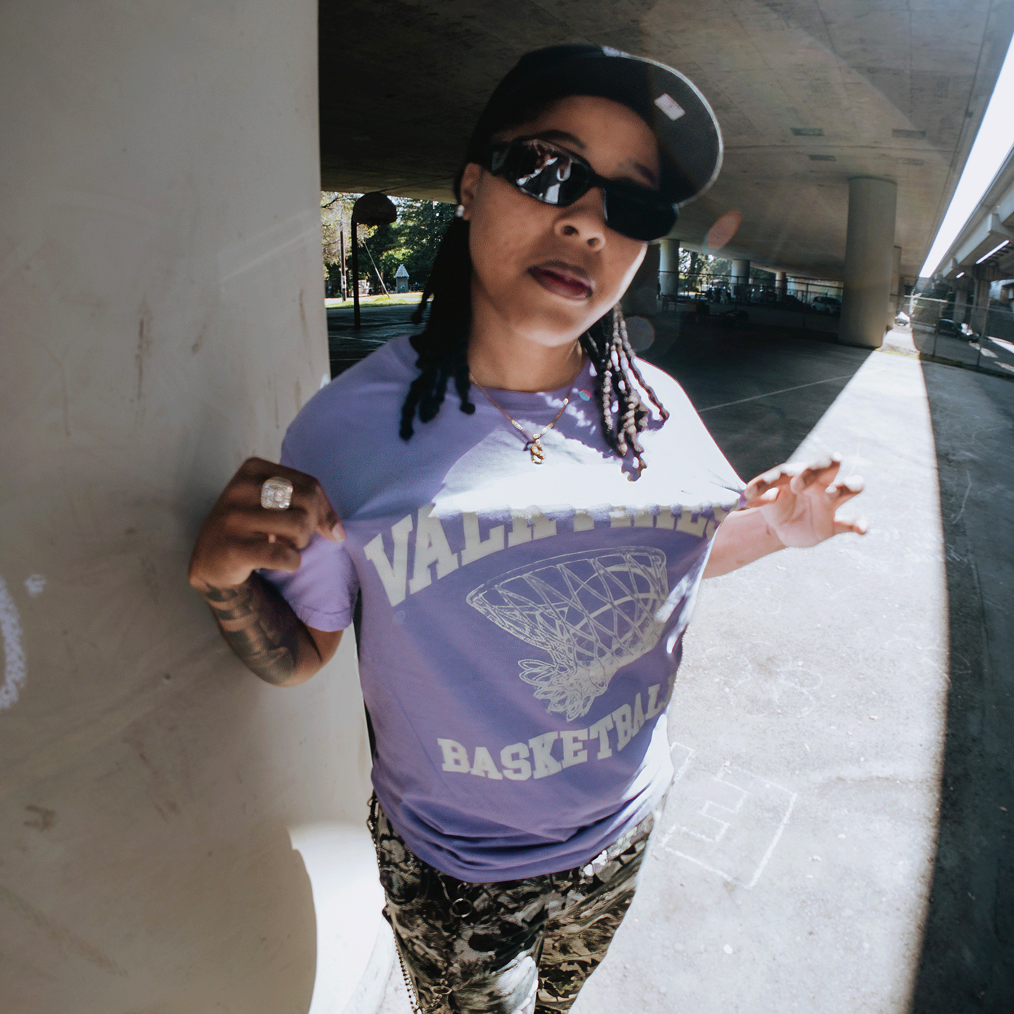 A person with long braids, sunglasses, and a baseball cap poses outdoors under a bridge, holding out the GSV Golden State Valkyries Rec League Premium Tee featuring a bold basketball net graphic and "VALID RUNS BASKETBALL" text.