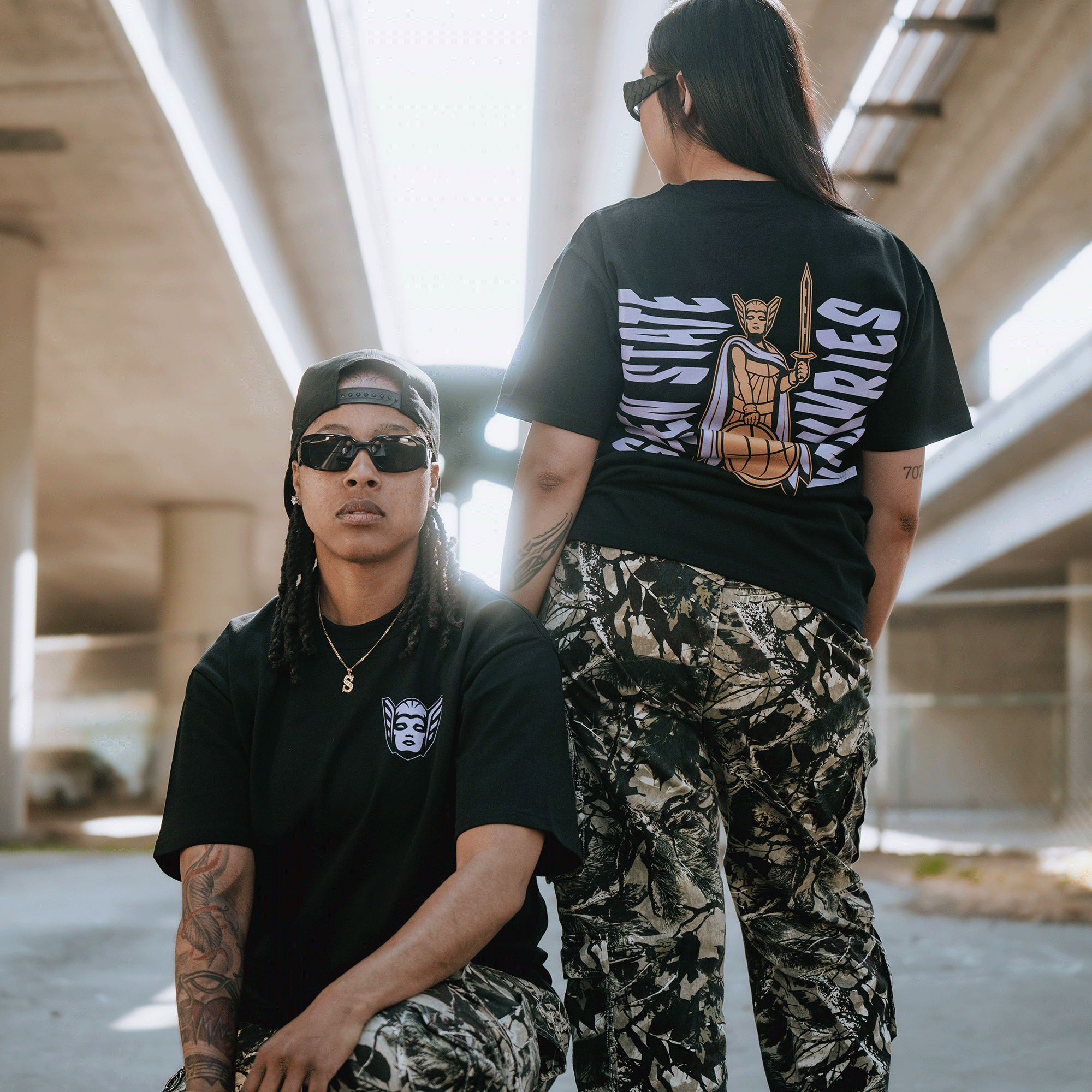 Two people in sunglasses and camouflage pants pose under a highway, exuding confidence. One stands, showcasing the GSV Golden State Valkyries Renowned Heavy Tee, featuring a bold basketball goddess graphic in an urban setting.