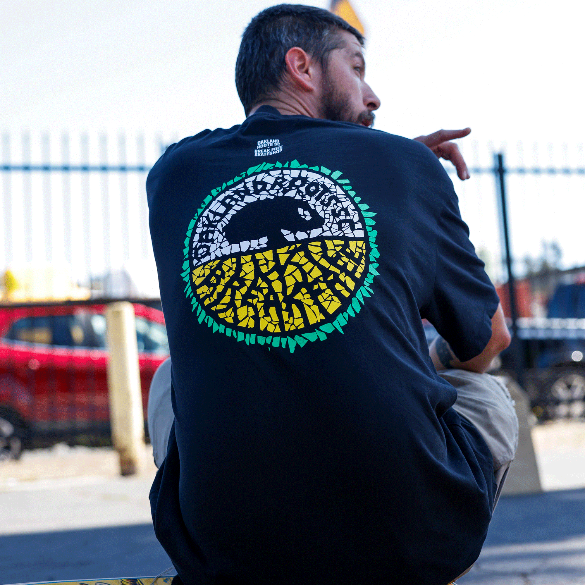 A man with short dark hair and a beard sits outdoors, facing away. He wears the Oakland Roots SC X Break Free Tee, featuring a colorful mosaic design inspired by Oakland culture. A fence and red car are blurred in the background.