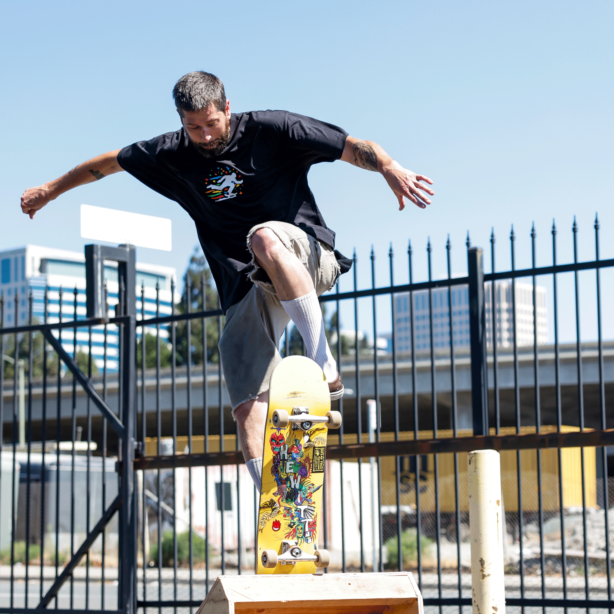 Wearing the Oakland Roots SC X Break Free Tee, a tattooed man skateboards over a wooden ramp. A black metal fence, city buildings, and blue sky set the scene—showcasing vibrant Oakland culture and style from Oakland Roots SC.