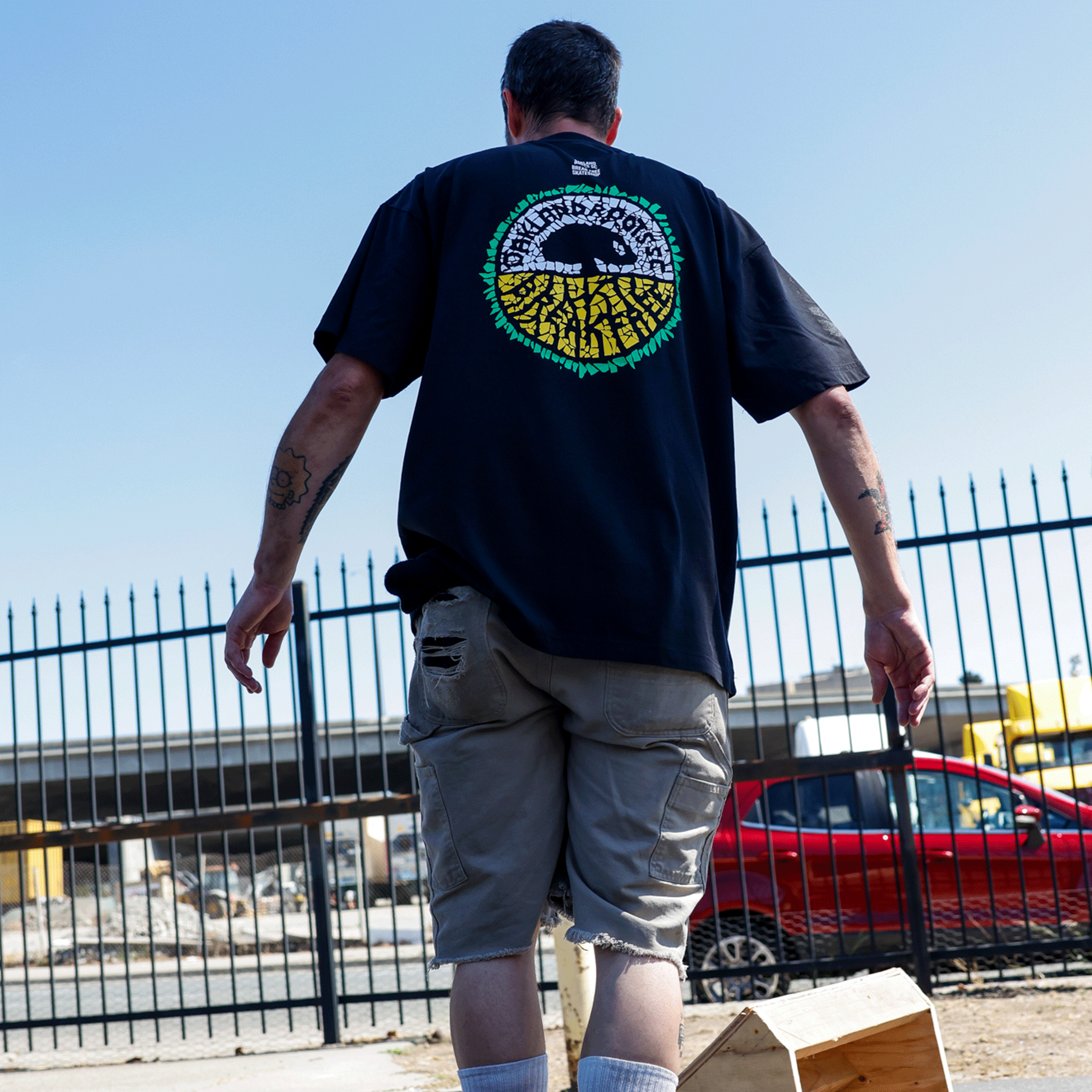 Wearing the Oakland Roots SC X Break Free Tee, a man stands outdoors near a black iron fence, showing off his tattoos. He embraces Oakland culture as he faces away from the camera, with a red car and city buildings behind him.