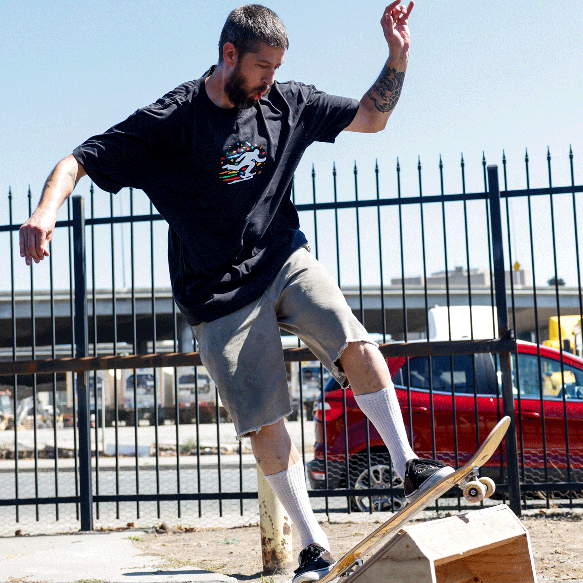 A tattooed, bearded man skates outdoors in Oakland culture style wearing the Oakland Roots SC X Break Free Tee by Oakland Roots SC, with gray shorts, white socks, and black shoes. Urban scenery, a red car, and a black fence appear under a clear blue sky.