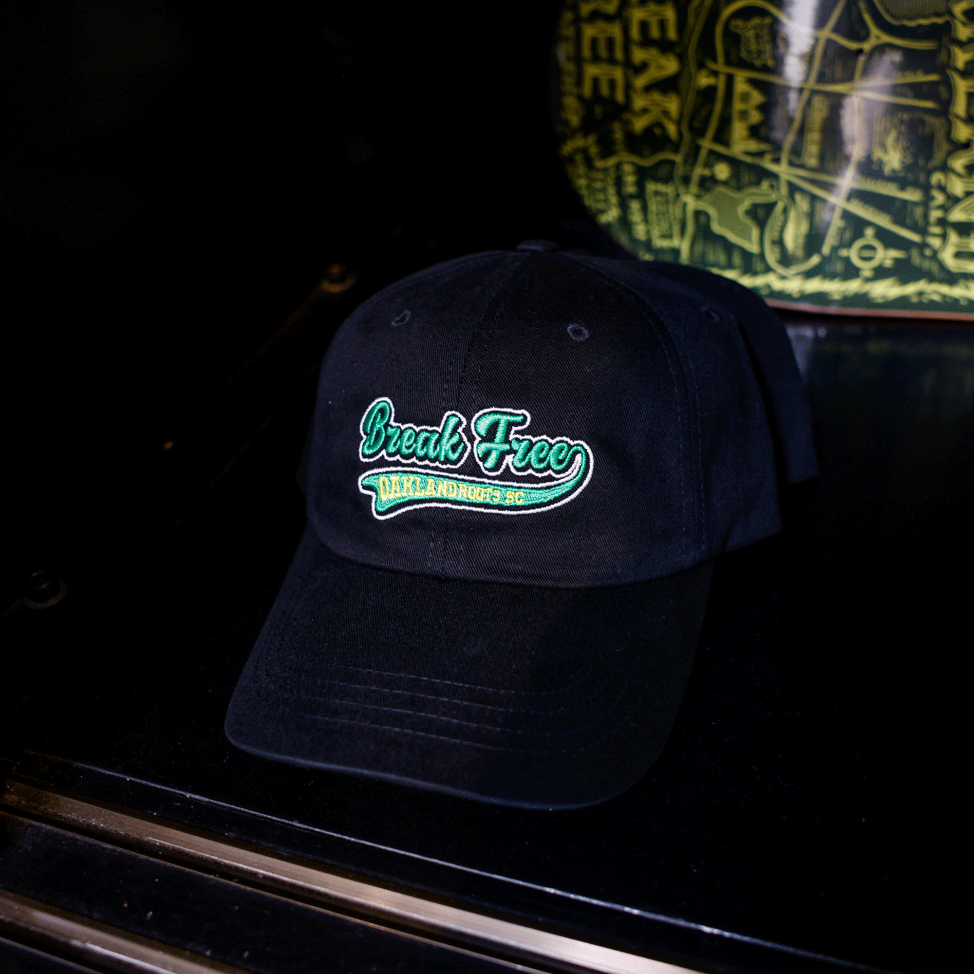 An Oakland Roots SC X Break Free Hat in black, featuring green and white "Break Free" embroidery with yellow accents, rests on a dark surface. A yellow-printed Oakland Roots SC item appears in the background.
