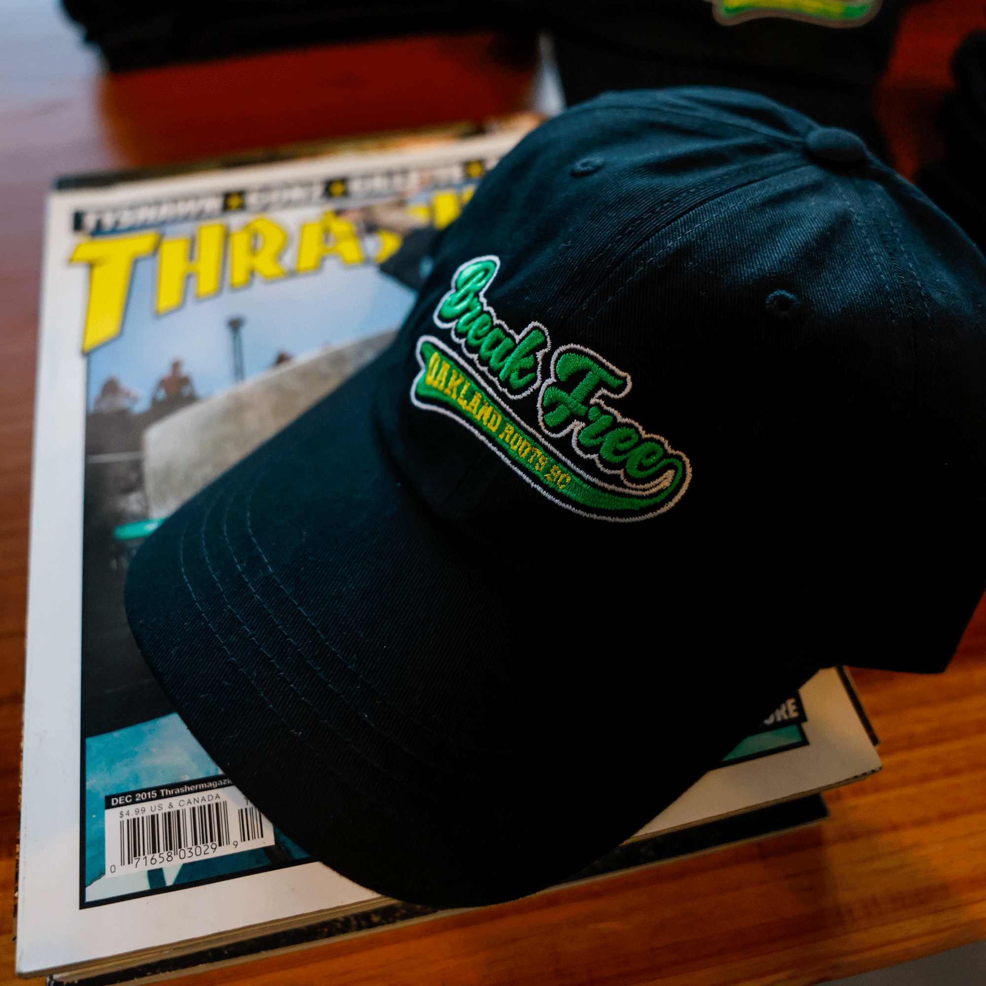 The Oakland Roots SC X Break Free Hat, a black dad hat with green and white embroidery, rests on a Thrasher skateboarding magazine atop a wooden surface. Its curved bill faces the viewer. Brand: Oakland Roots SC.