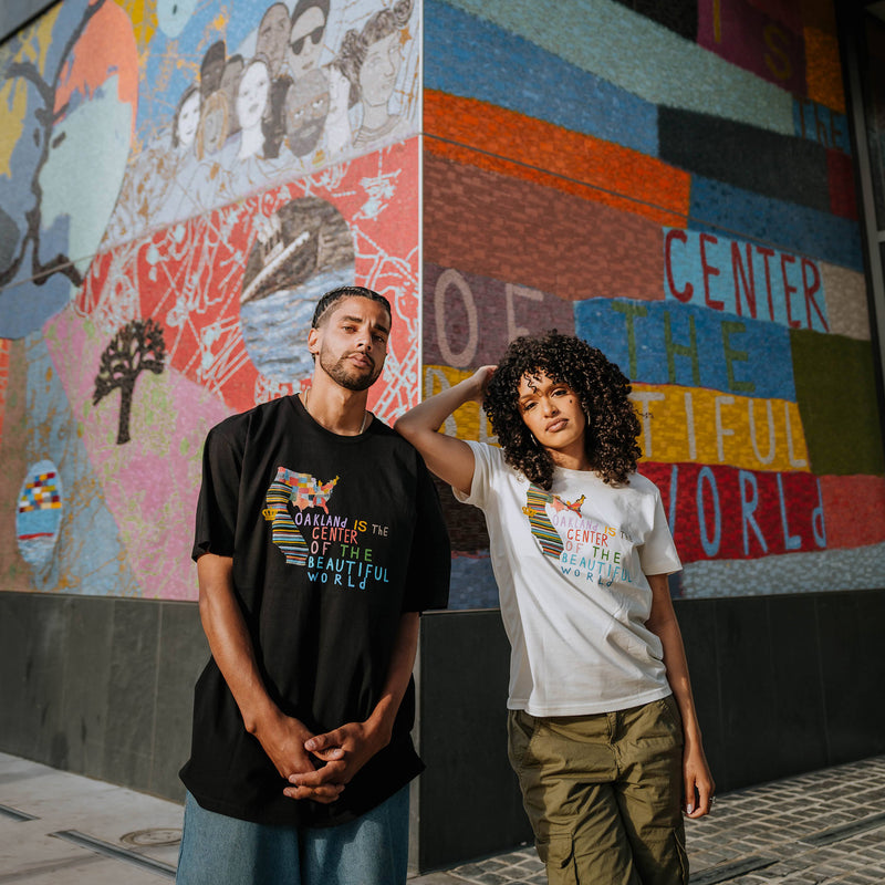 Male and Female models wearing Oakland Is the Center of World mural art by contemporary artist Squeak Carnwath on a black & natural colored t-shirt.