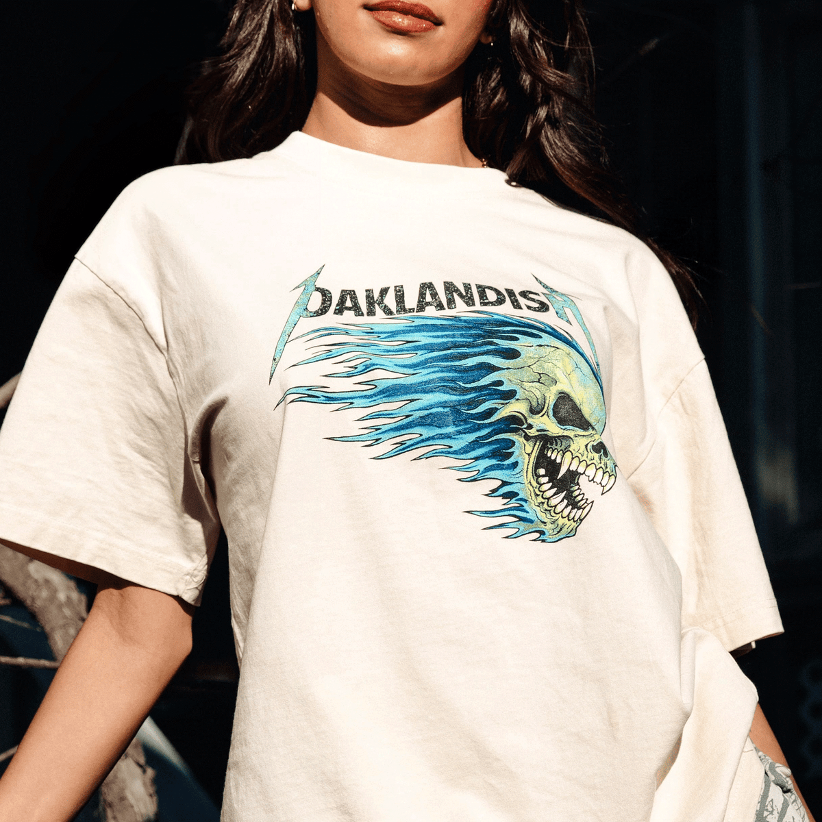 A person wears a cream-colored, classic fit Sandman Tee by Oaklandish, featuring a yellow skull in blue flames and the brand name in a metallic font, set against bright lighting that casts shadows behind the subject.