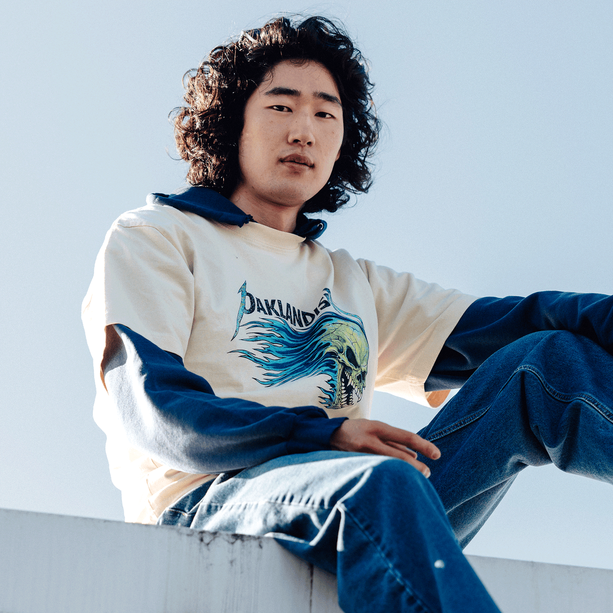 A person with shoulder-length dark curly hair sits outdoors against a clear blue sky, wearing an Oaklandish Sandman Tee—a cream-colored long-sleeve shirt with a blue hood and a graphic of a blue flaming skull on the chest—paired with blue jeans.