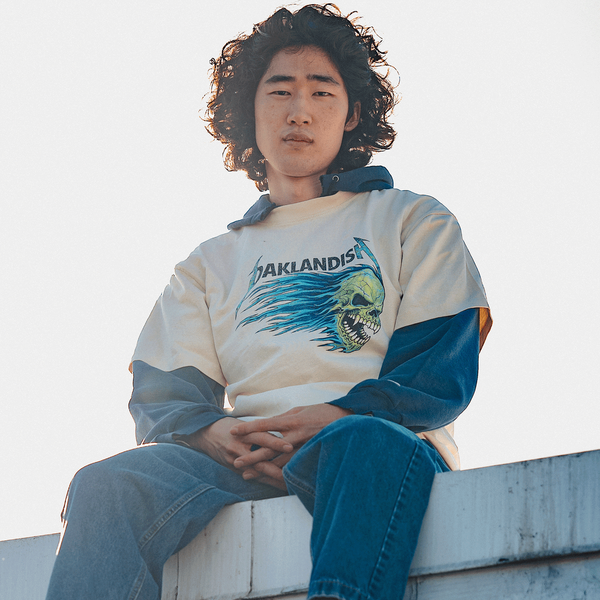 A young person with shoulder-length curly hair sits on a ledge against a hazy sky, wearing an Oaklandish Sandman Tee over a navy hoodie and blue jeans, looking at the camera with hands clasped.