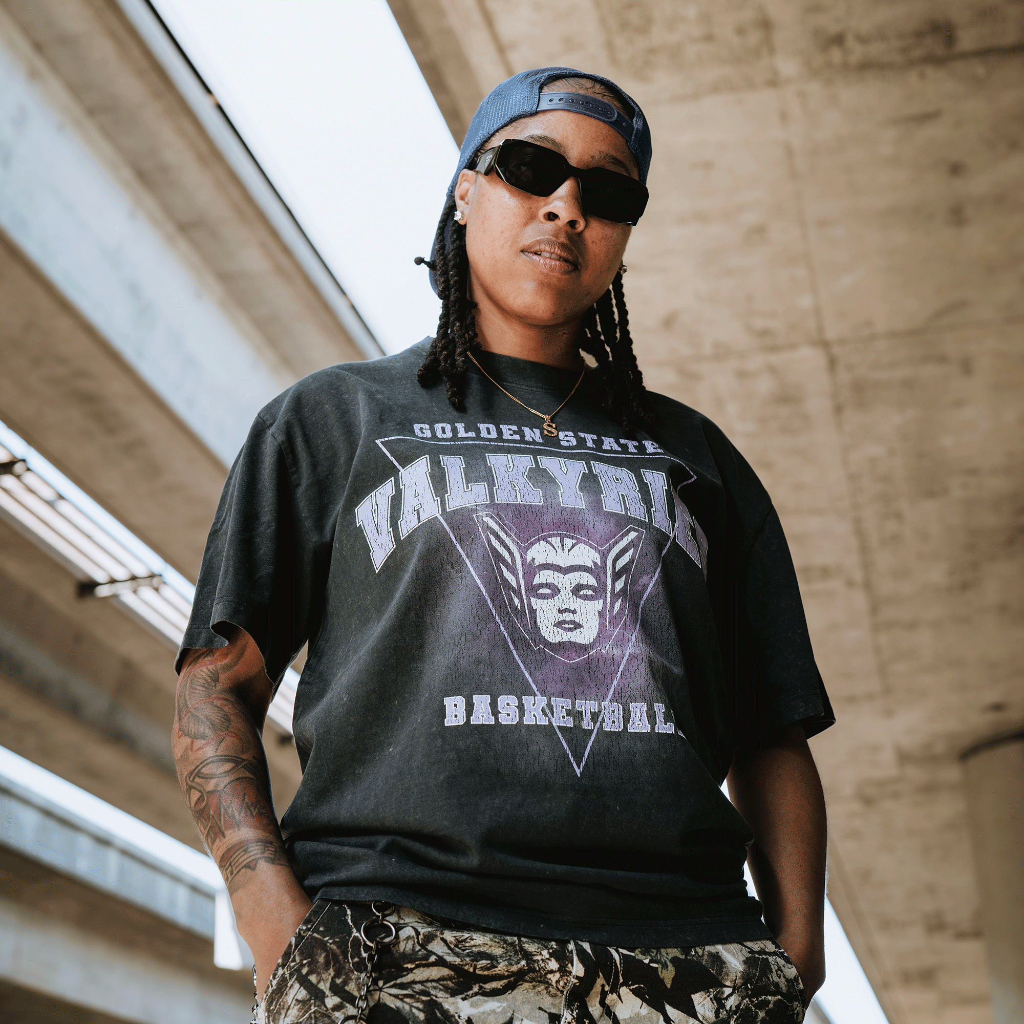 A person with long braids, sunglasses, and a cap stands under an overpass. They wear the GSV Golden State Valkyries Surge Stone Wash Heavy Tee and camouflage pants, exuding confidence with tattoos on one arm and hands in pockets.