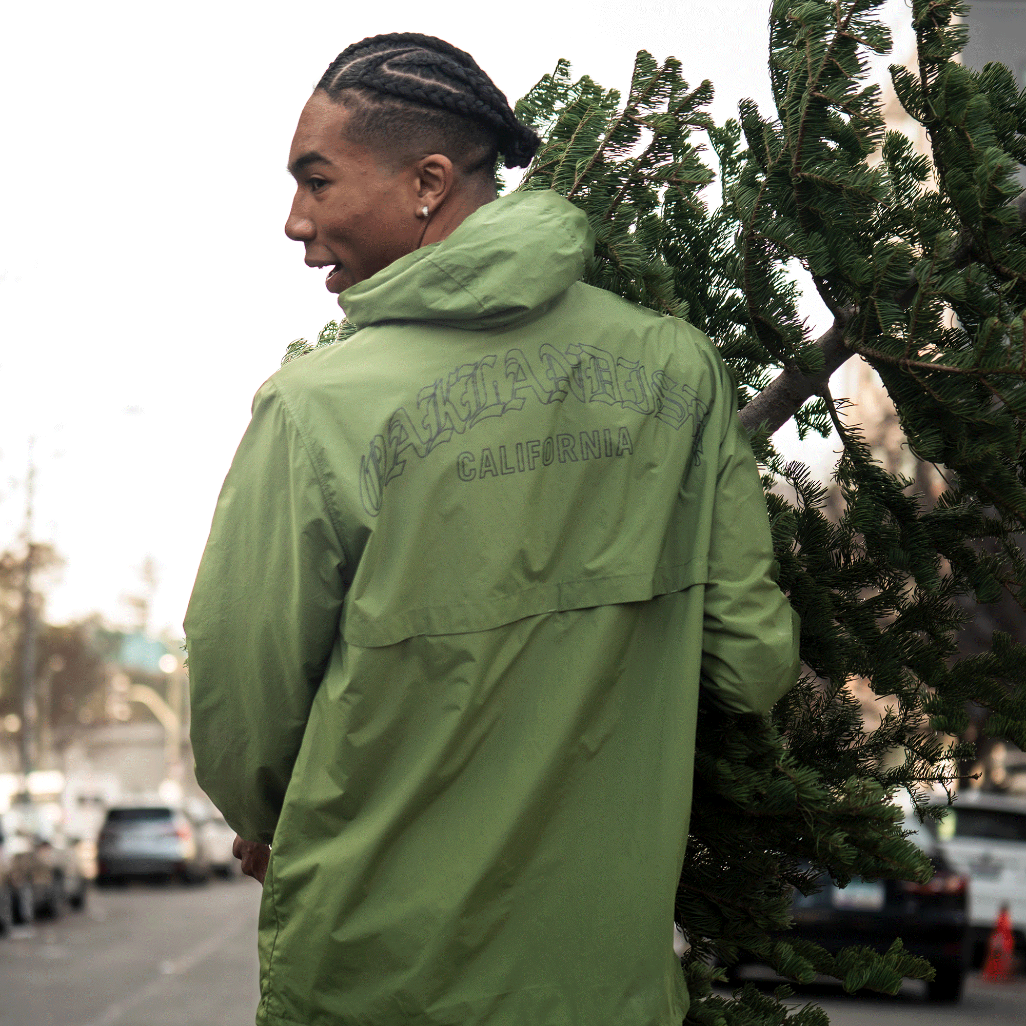 Wearing a classic fit green Oaklandish Susurrus Track Top, a person smiles as they carry a Christmas tree over their shoulder on a city street lined with trees and parked cars, looking back in the daytime.