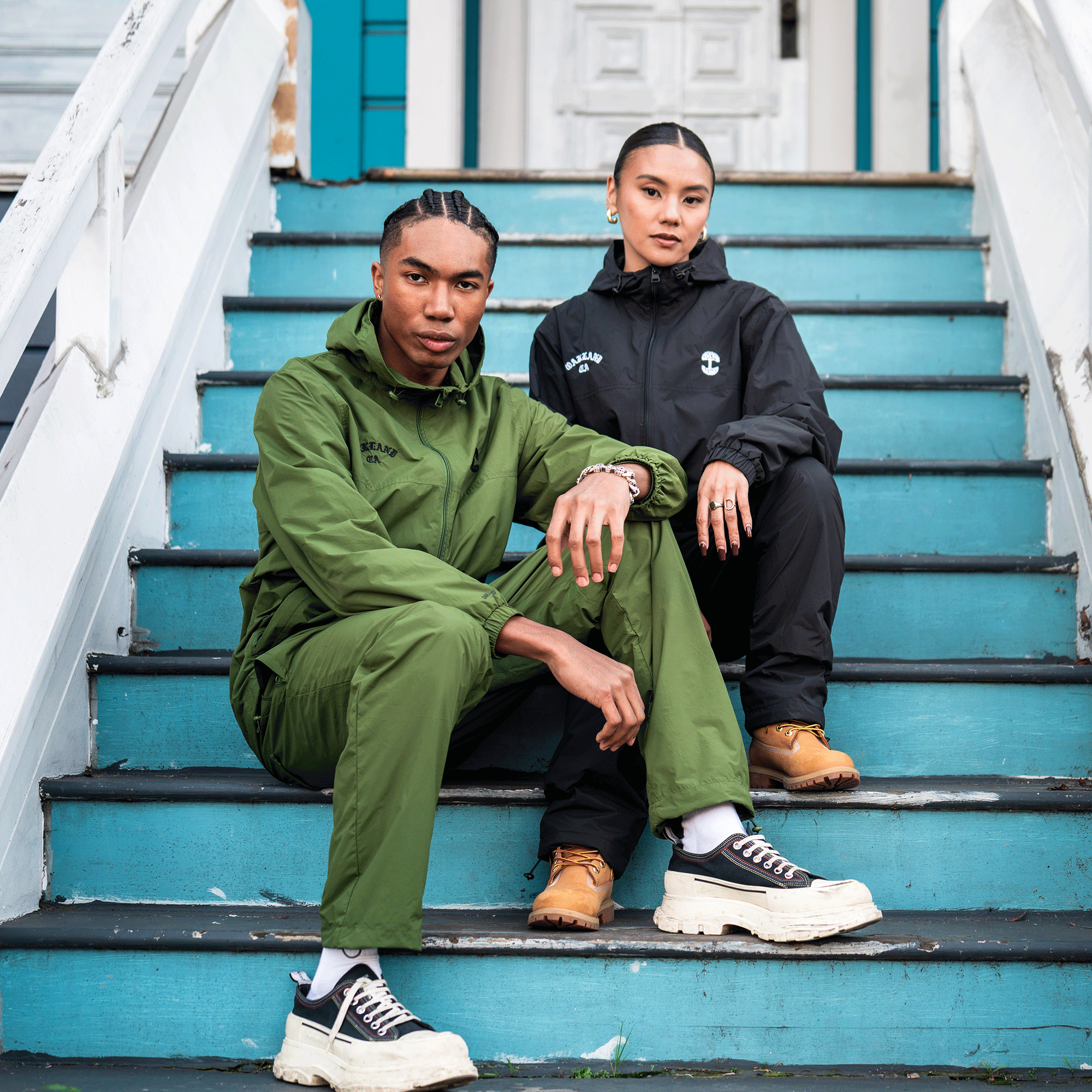 Two people sit on turquoise outdoor stairs. The person on the left wears a green tracksuit with an embroidered tree logo, white socks, and sneakers. The person on the right wears Oaklandish Susurrus Track Bottoms and tan boots.