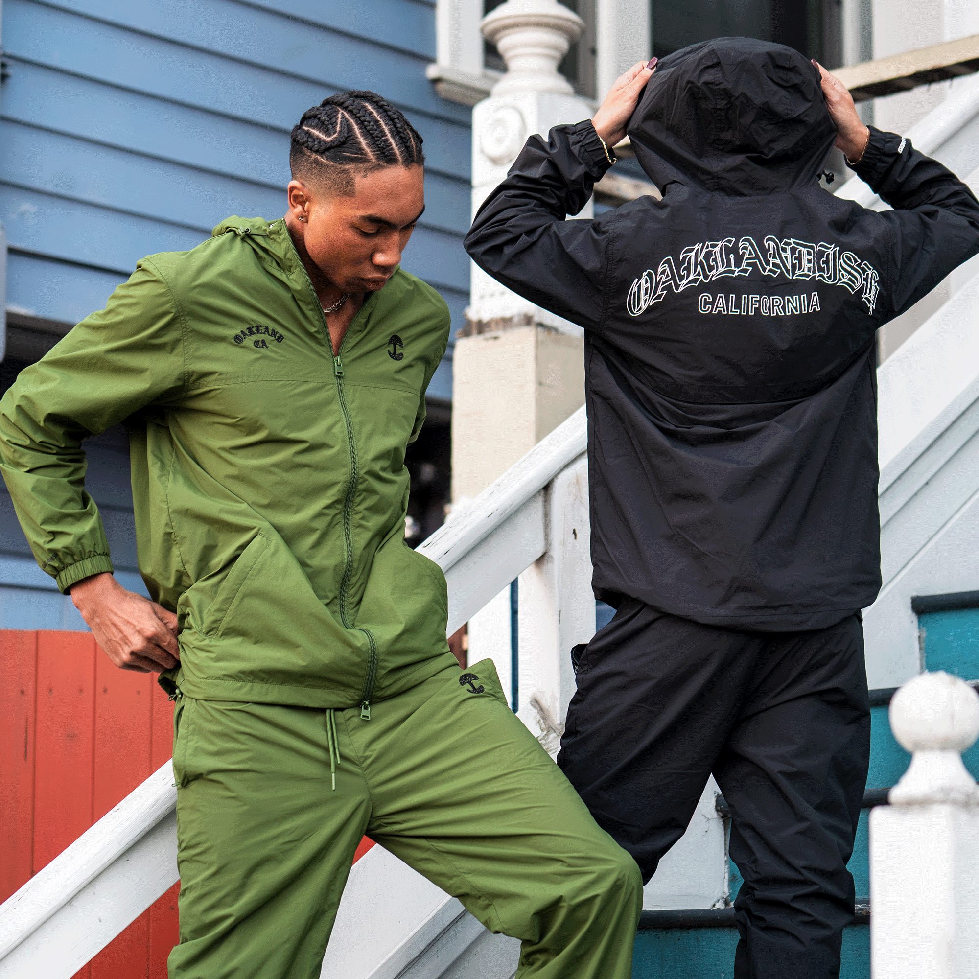 Two people in sporty tracksuits stand on outdoor stairs. One wears a green "OAKLANDISH" top, facing forward. The other adjusts their hood, wearing Oaklandish Susurrus Track Bottoms—black nylon pants with an adjustable waistband and logo on the back.