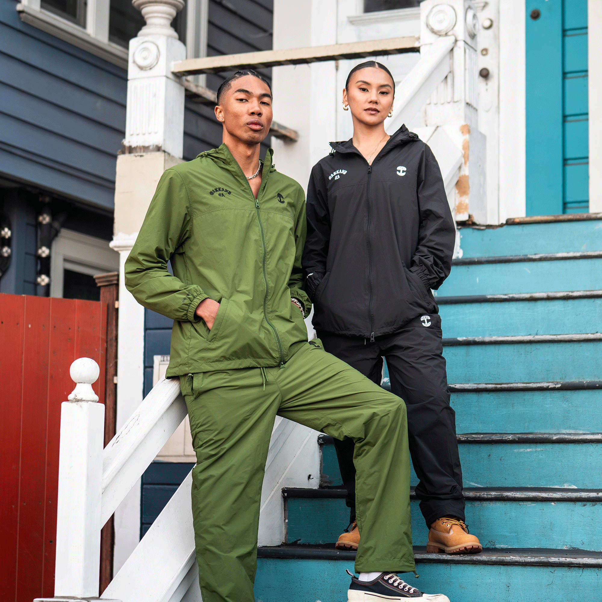 Two people pose on blue steps outside a house. The person on the left wears the Oaklandish Susurrus Track Top and matching bottoms, while the person on the right sports a black hooded nylon zip jacket, black pants, and tan boots. Both look confidently at the camera.