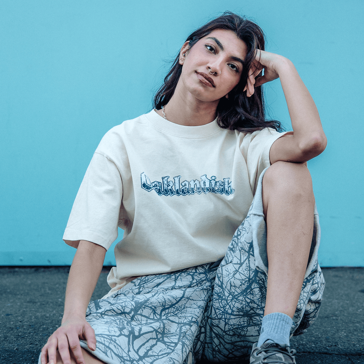 A young person with long dark hair and a septum piercing sits against a bright blue wall, wearing an Oaklandish Swoop Tee with a blue chest graphic and patterned grey shorts with a tree-branch print.