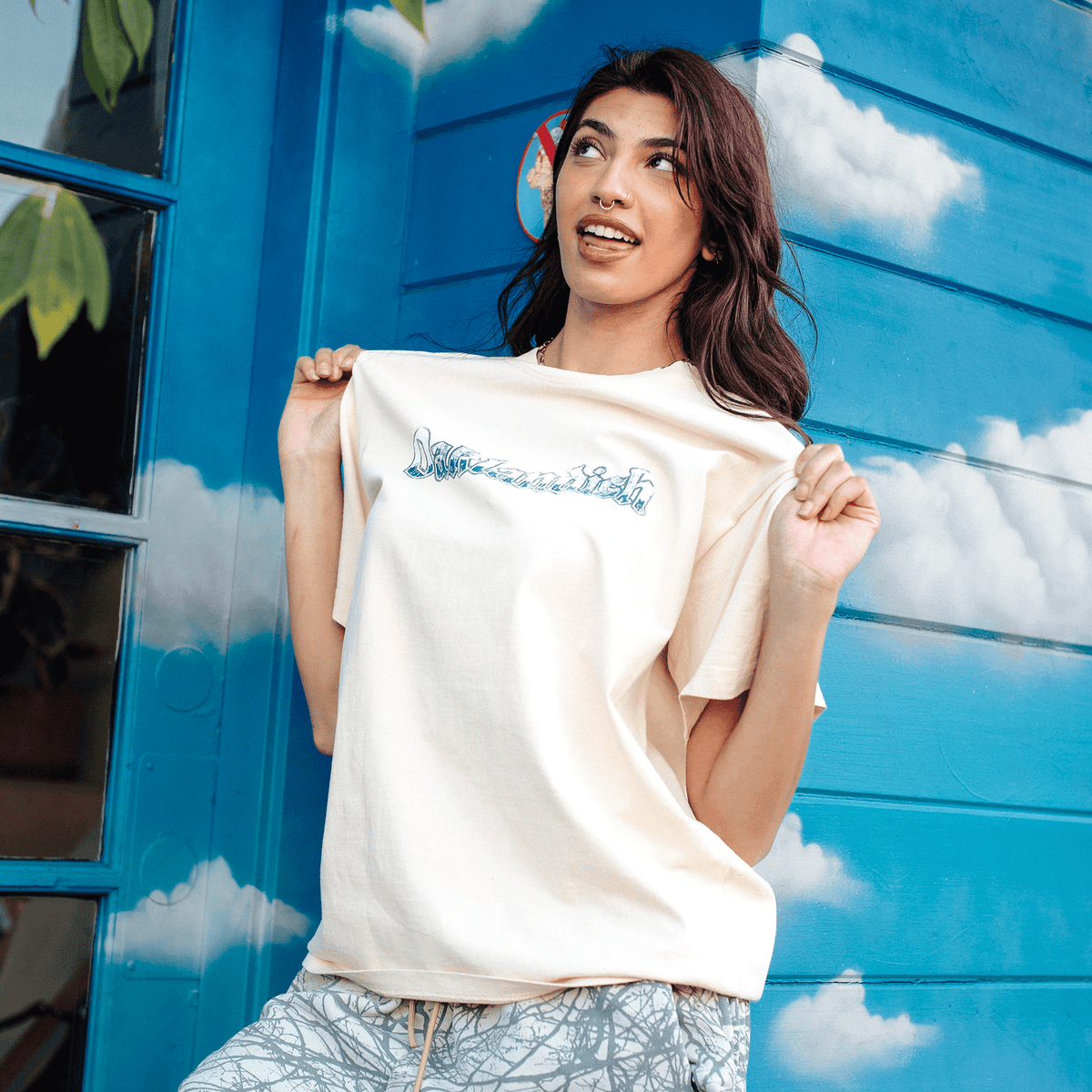 A woman with long brown hair and a septum piercing poses in an Oaklandish Swoop Tee made of heavy-weight cotton. She pulls the cream-colored fabric with blue cursive text taut against a blue wall painted with white clouds, wearing patterned gray trousers while looking to the side.
