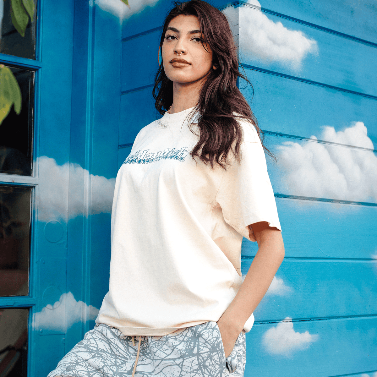 A person with long, dark wavy hair and a septum piercing poses in a cream-colored Oaklandish Swoop Tee with blue lettering, wearing patterned drawstring pants while standing against a bright blue wall painted with white clouds.