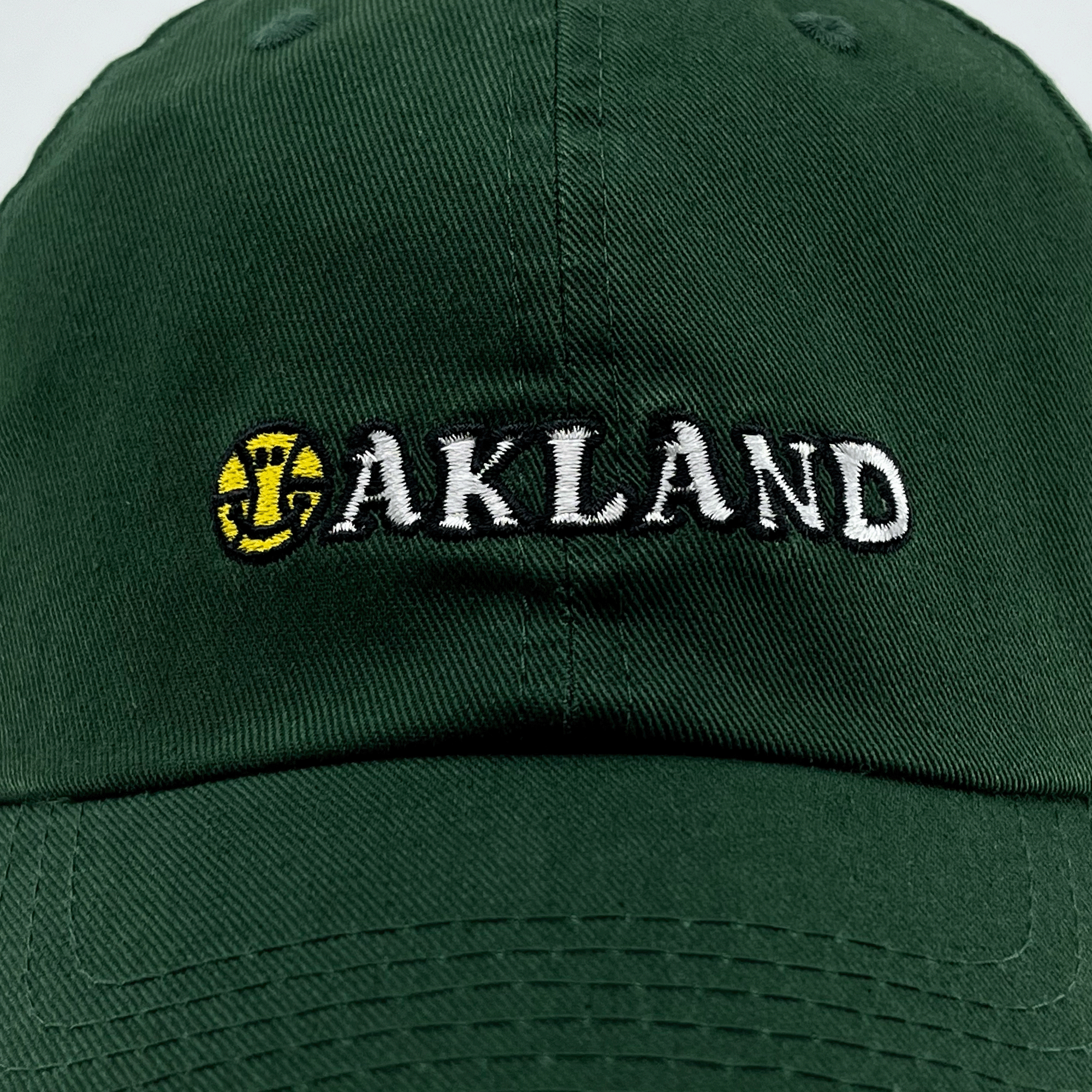 The Happy Oakland Dad Hat by Oaklandish features a dark green color with "OAKLAND" embroidered on the front in white, uppercase letters. The "O" is uniquely designed as a yellow baseball with red seams and an overlapping green-colored bat emblem. This cap embodies Bay Area culture with its visible stitching and a curved brim.