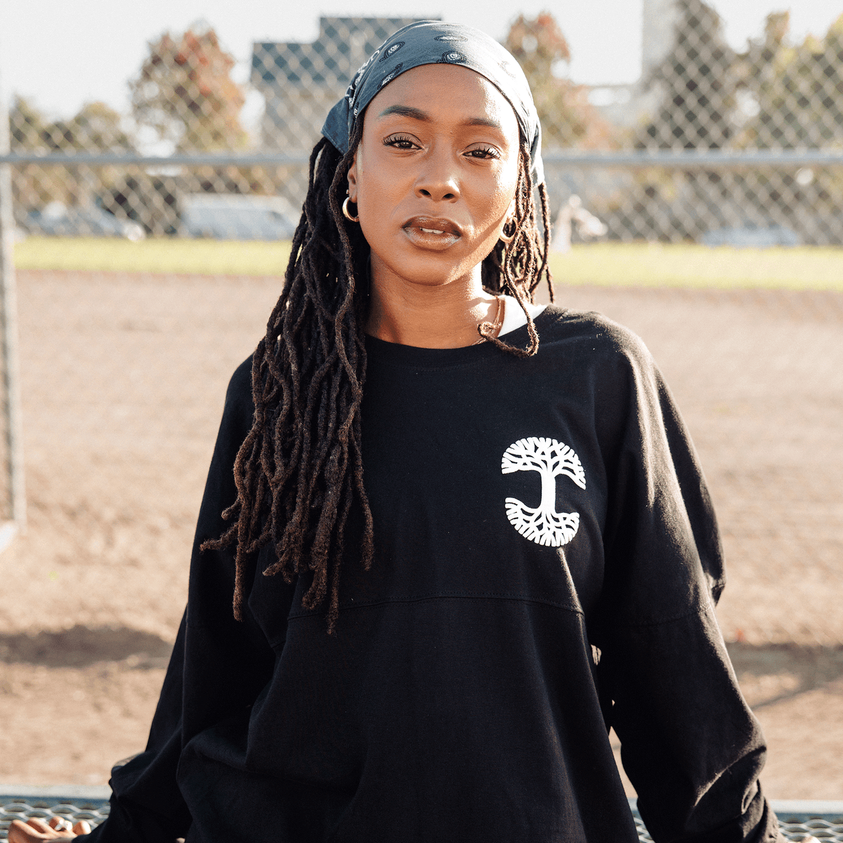 A woman with long dreadlocks and a dark patterned bandana poses in front of a chain-link fence, wearing an Oaklandish Temple LS Tee with a white circular tree graphic on the chest in a sunny outdoor field.