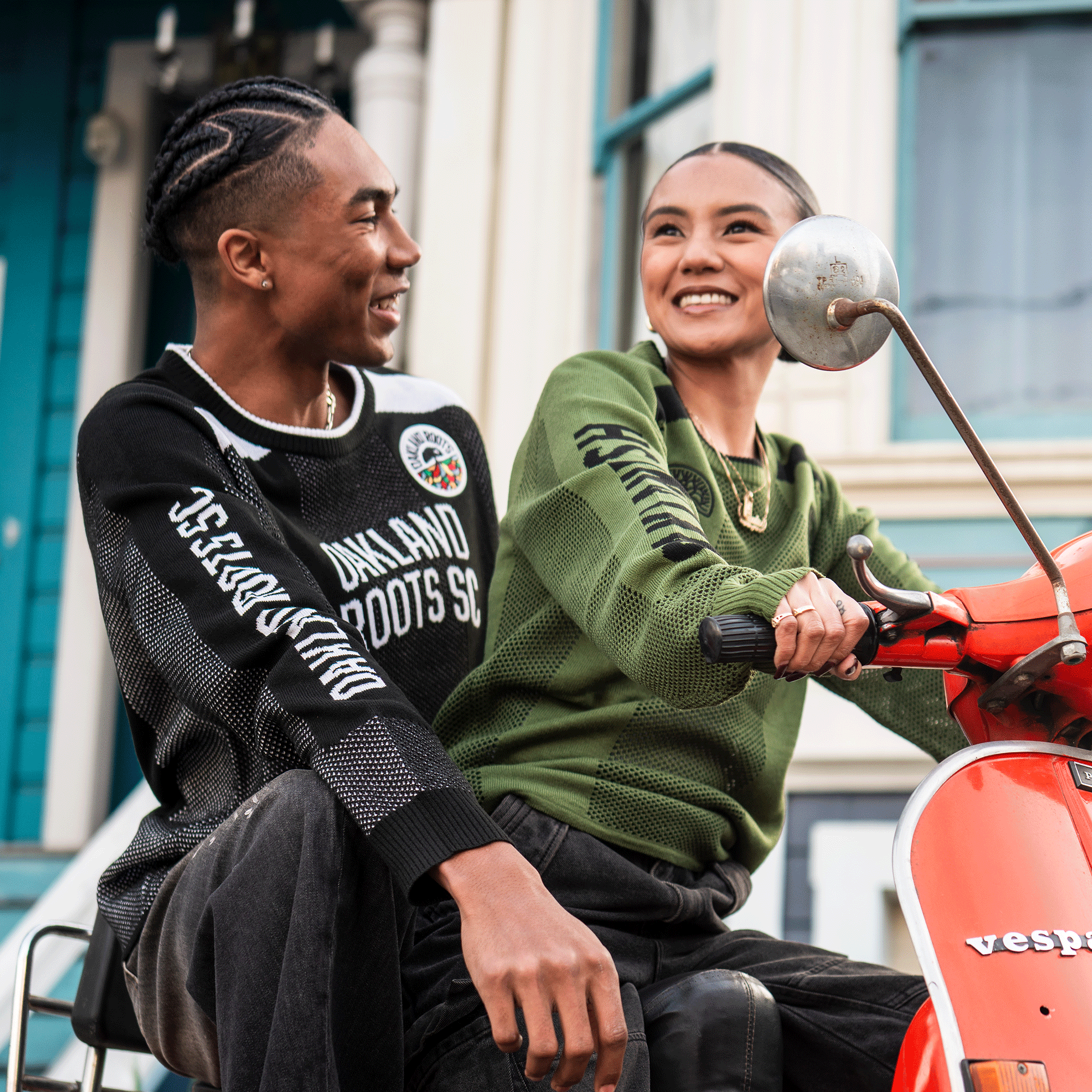 Two young people smile on a bright orange Vespa. The left wears a unisex Oakland Roots SC Clubhouse Sweater; the right, who’s driving, sports a green Oaklandish knit. They're outside near colorful buildings.