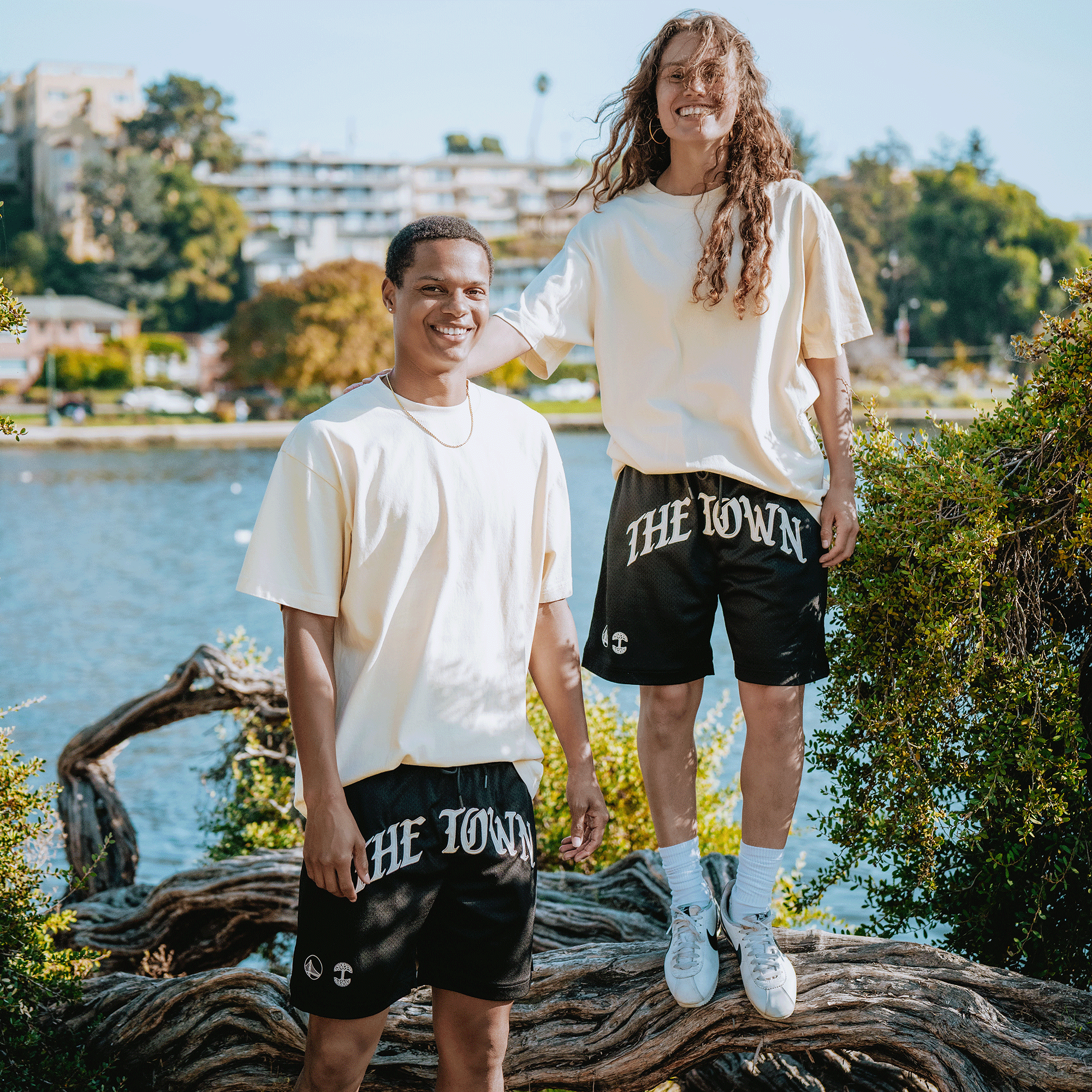 Two smiling people stand by a lake in greenery, both wearing oversized white shirts and GSW Arched Black-Letter Shorts. One is on a log while the other stands beside it, with buildings and trees visible in the background.