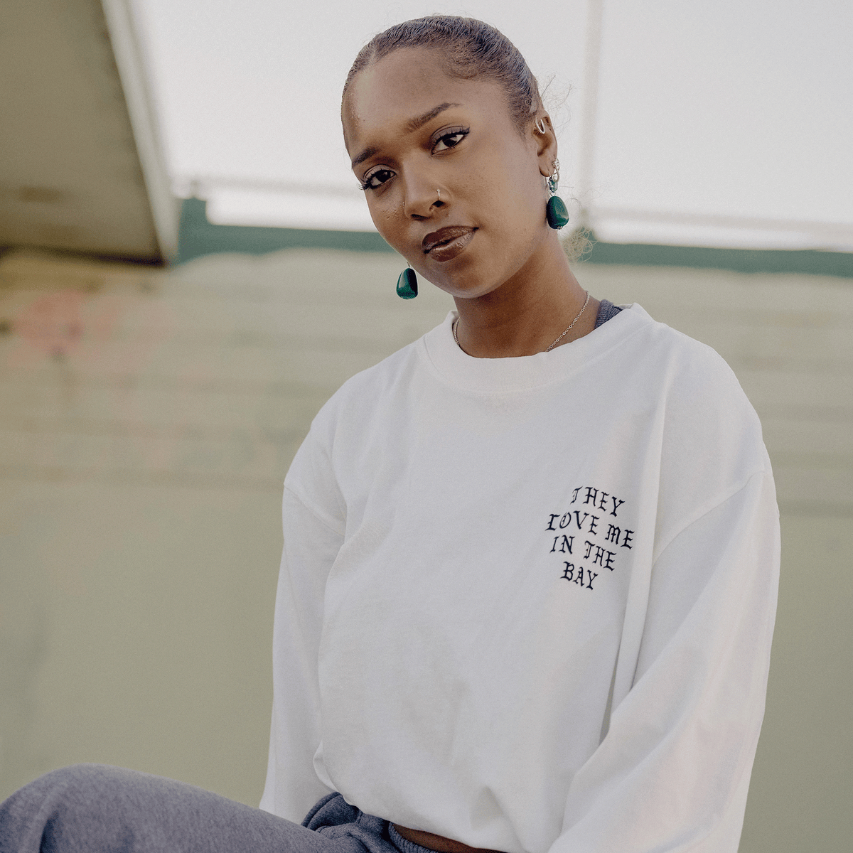 A woman with dark hair pulled back looks at the camera, wearing a They Love Me LS Tee By Doc in white with black text. She has blue drop earrings and is posed outdoors against a muted background.