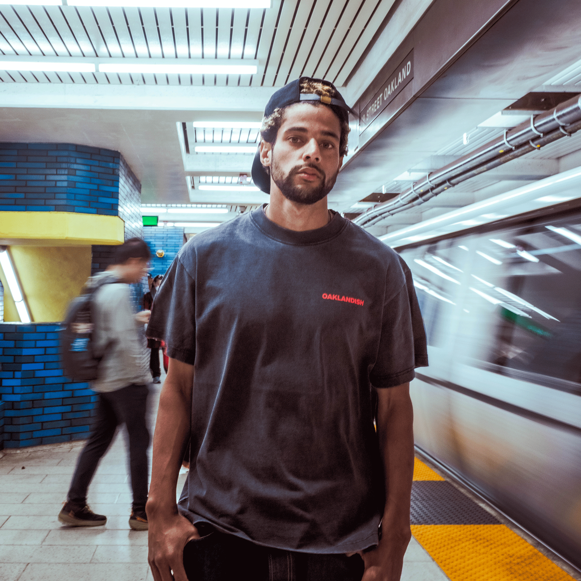 A young man in the Oaklandish Tiger Blood Tee and a backwards cap stands on a subway platform as a train speeds by, capturing classic style in a busy urban scene with blue tiles and overhead lights.