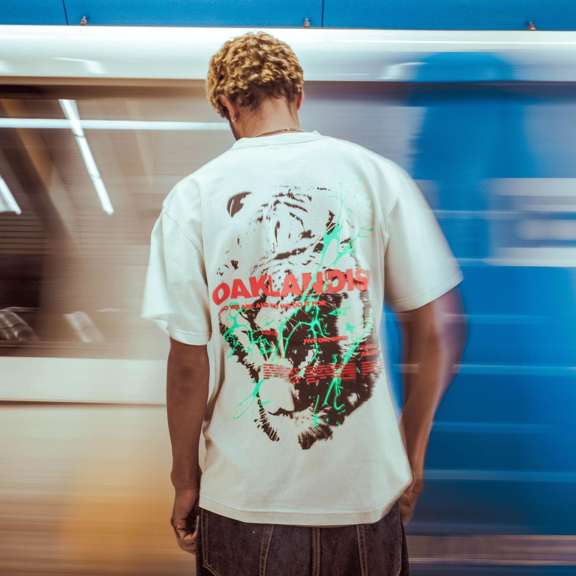A person with short, curly blond hair stands with their back to the camera, wearing the Oaklandish Tiger Blood Tee in white. "OAKLAND 015" is printed in red on the back as a blurred blue train moves by, adding a dynamic sense of motion.