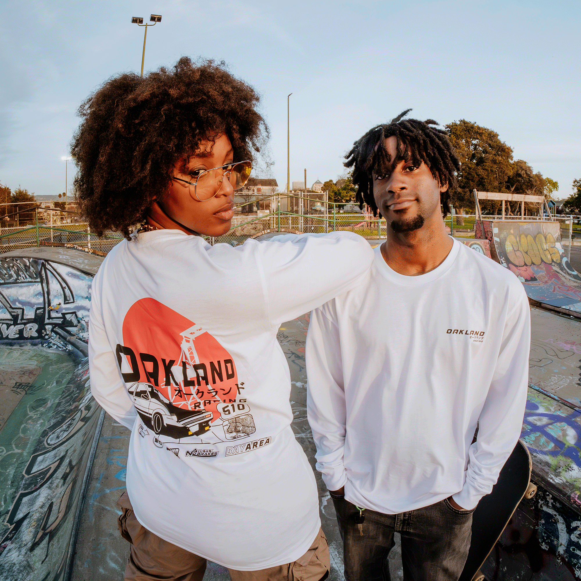 A person with curly hair and glasses faces away, wearing an Oaklandish Tuner LS Tee featuring graphic art on the back. Nearby, another with short dreadlocks faces forward in a matching 100% cotton shirt. They're in a graffiti-covered skate park under a blue sky.