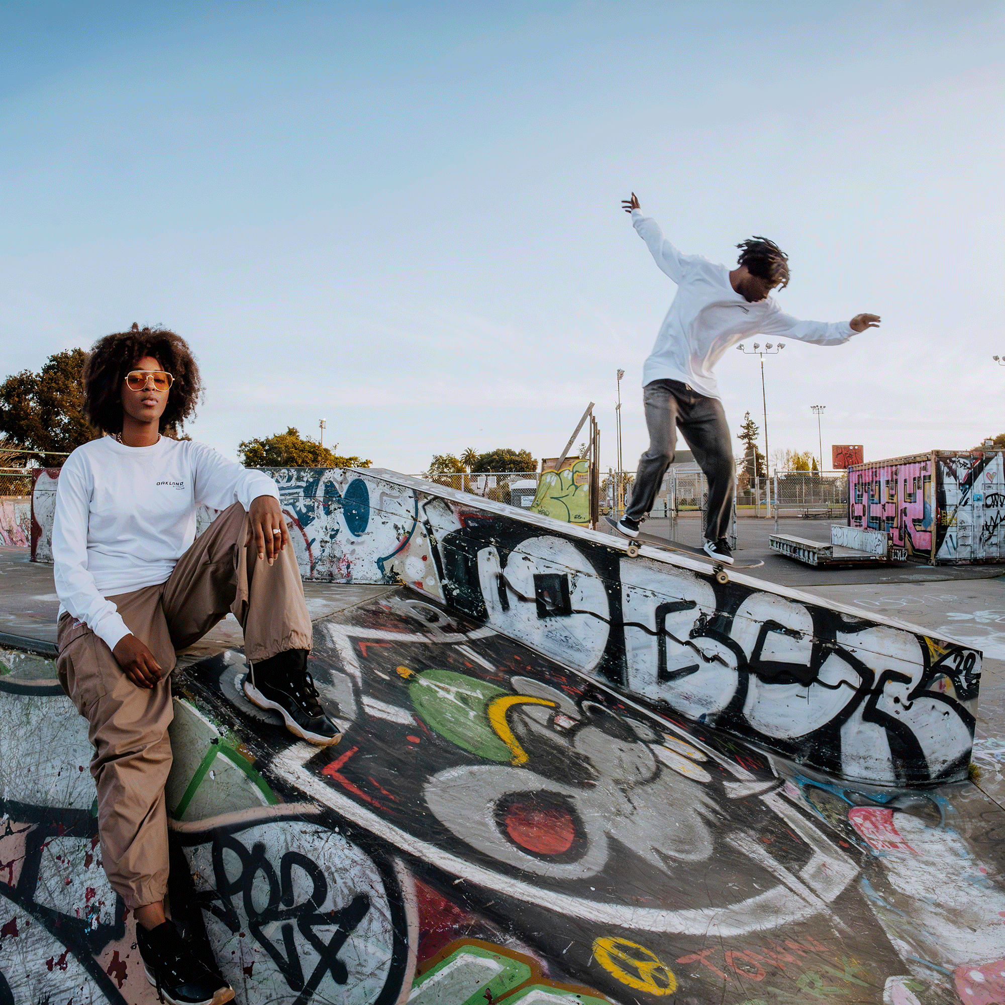 Sporting an Oaklandish Tuner LS Tee, a person sits on a graffiti-covered ramp at an open skate park, watching another skateboarder in white and brown navigate a slope under the clear sky.
