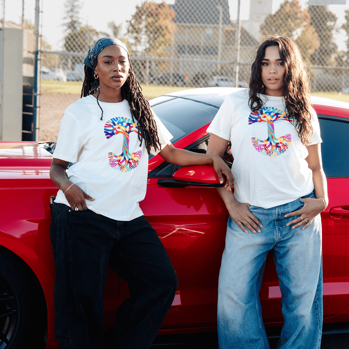 Two women wearing white Oaklandish Urban Tropics Tees pose in an outdoor parking lot in front of a bright red car; the woman on the left wears dark jeans and a headscarf, while the woman on the right wears light-wash wide-leg denim.